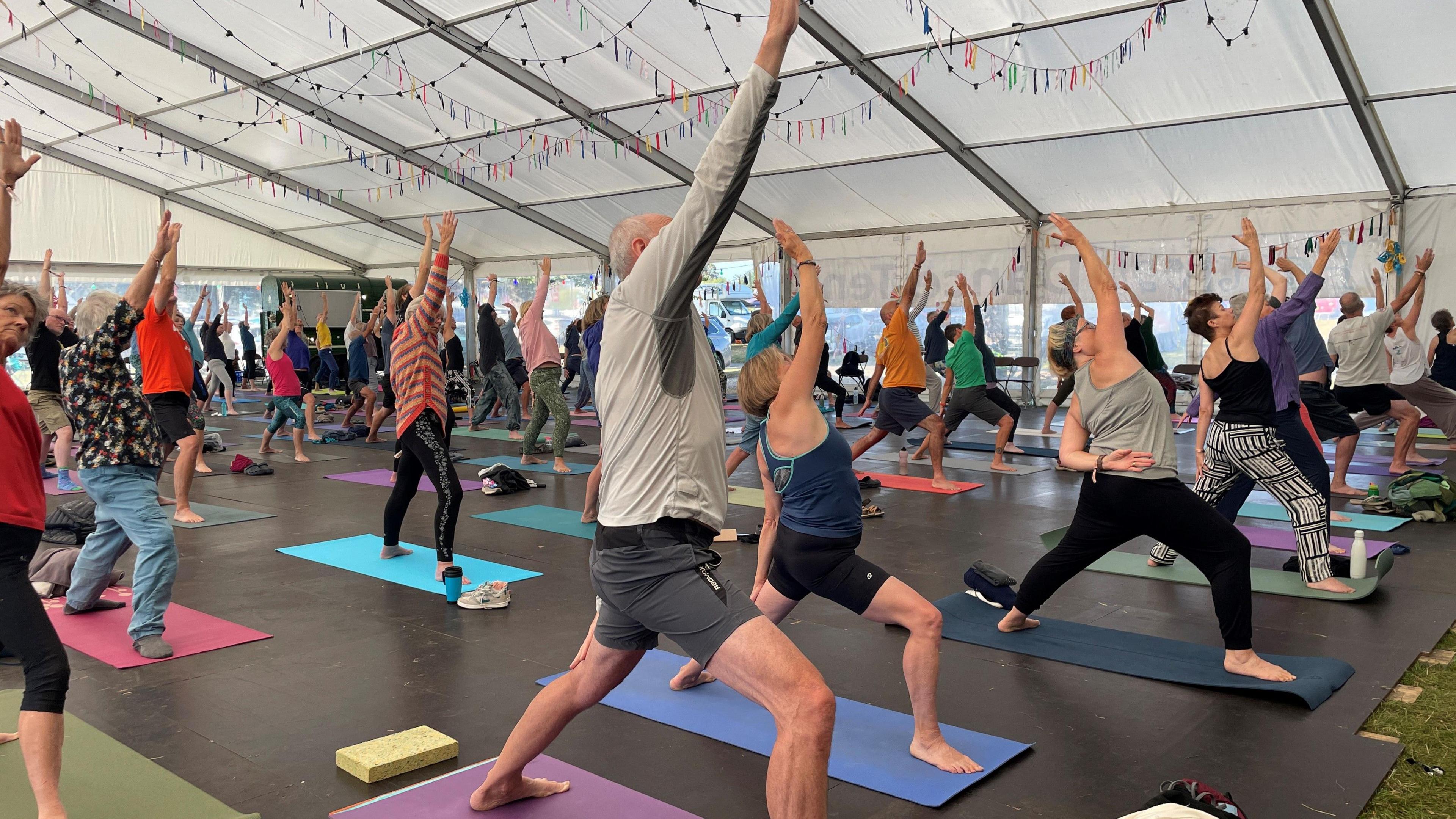 Men and women, taking part in a yoga workshop in a marquee. They are stepping forward in a long lunge and have one arm pointing straight up in the air, in the "reverse warrior" posture. They all have different colour mats that are laid on a black floor. 