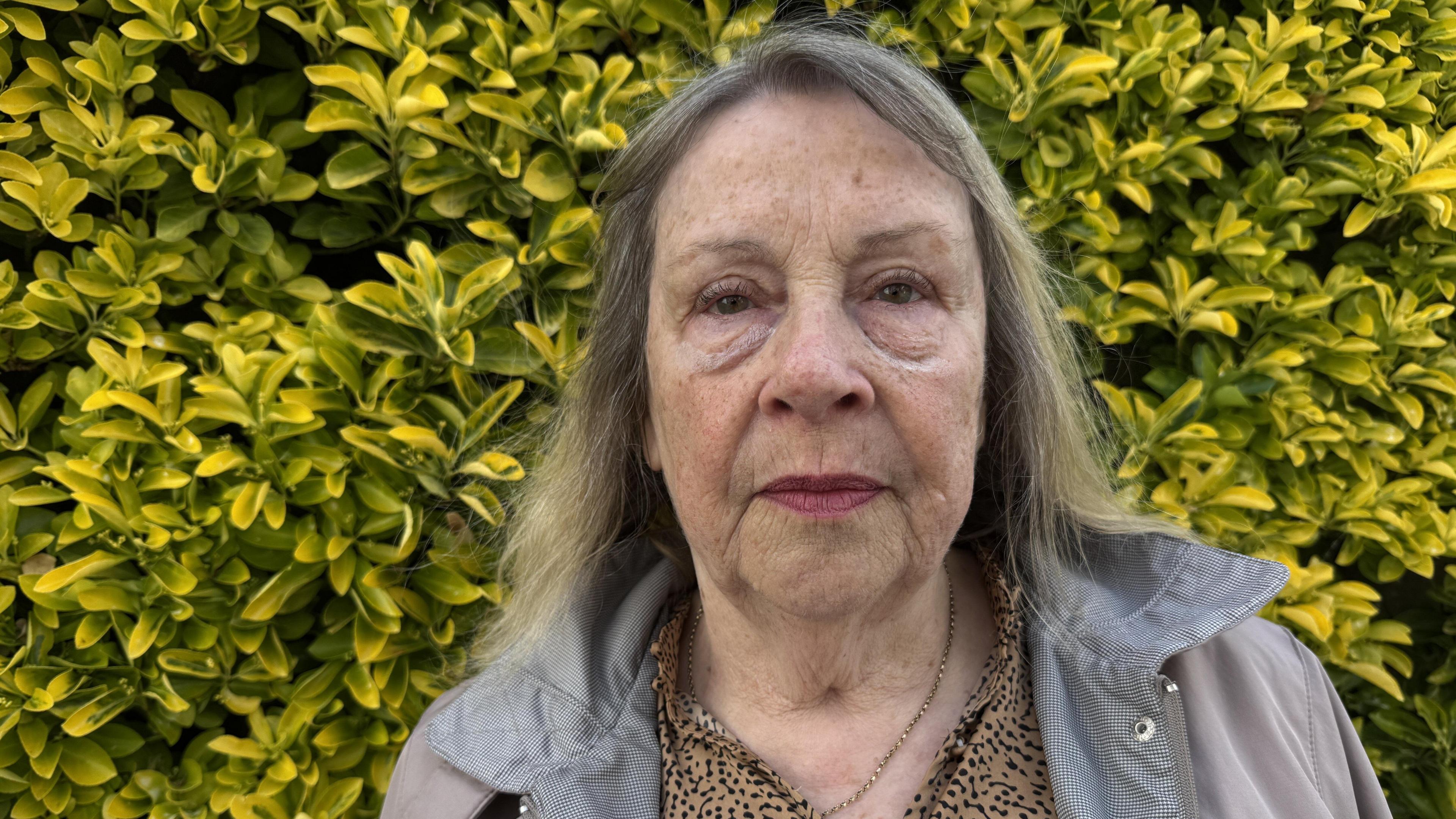 Woman in grey jacket stands in front of yellow/green hedge