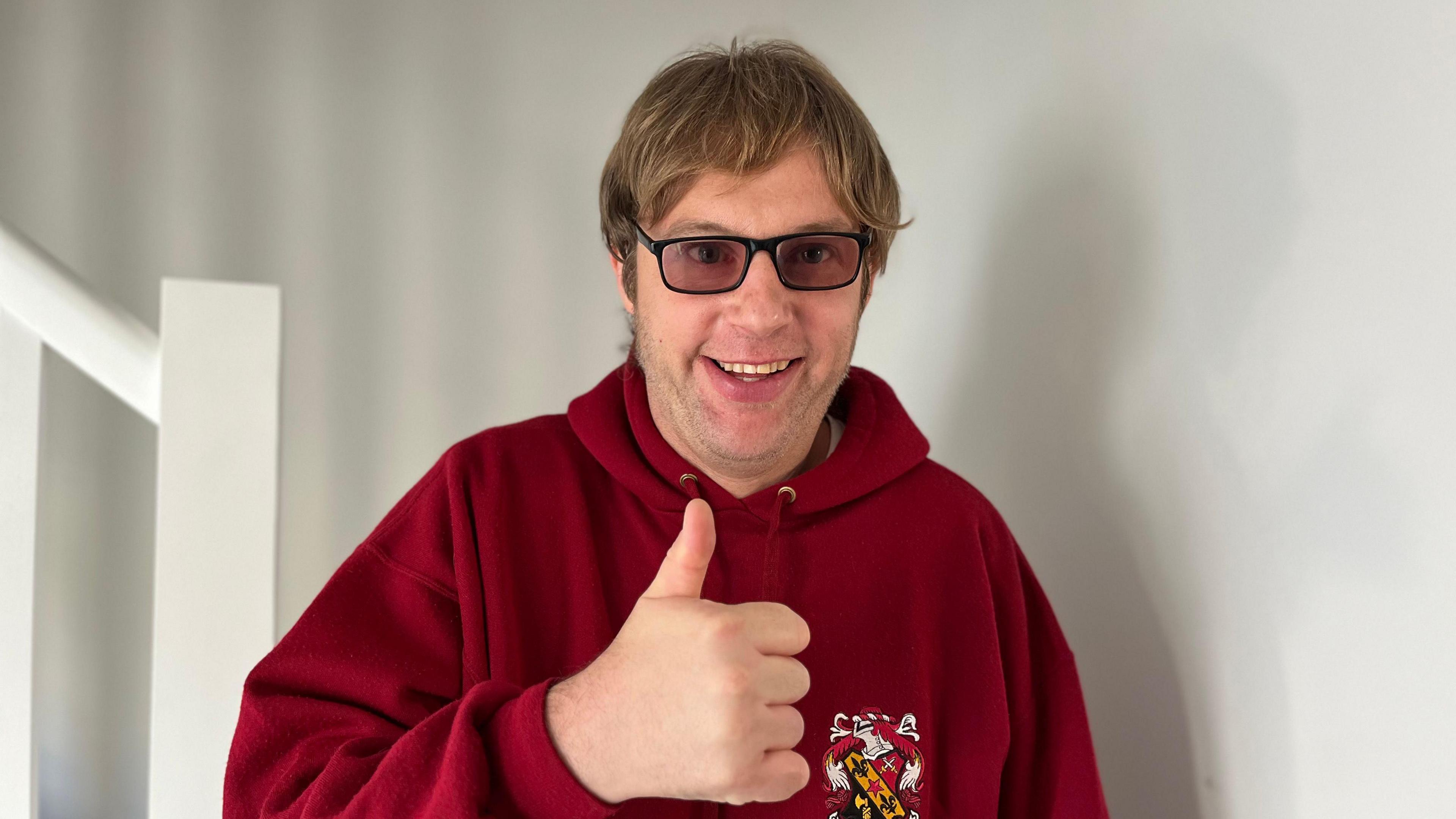 Martin wearing a red hooded jumper, with a badge on the right, against a white wall. He is putting his thumbs up to the camera and is wearing black tinted glasses.