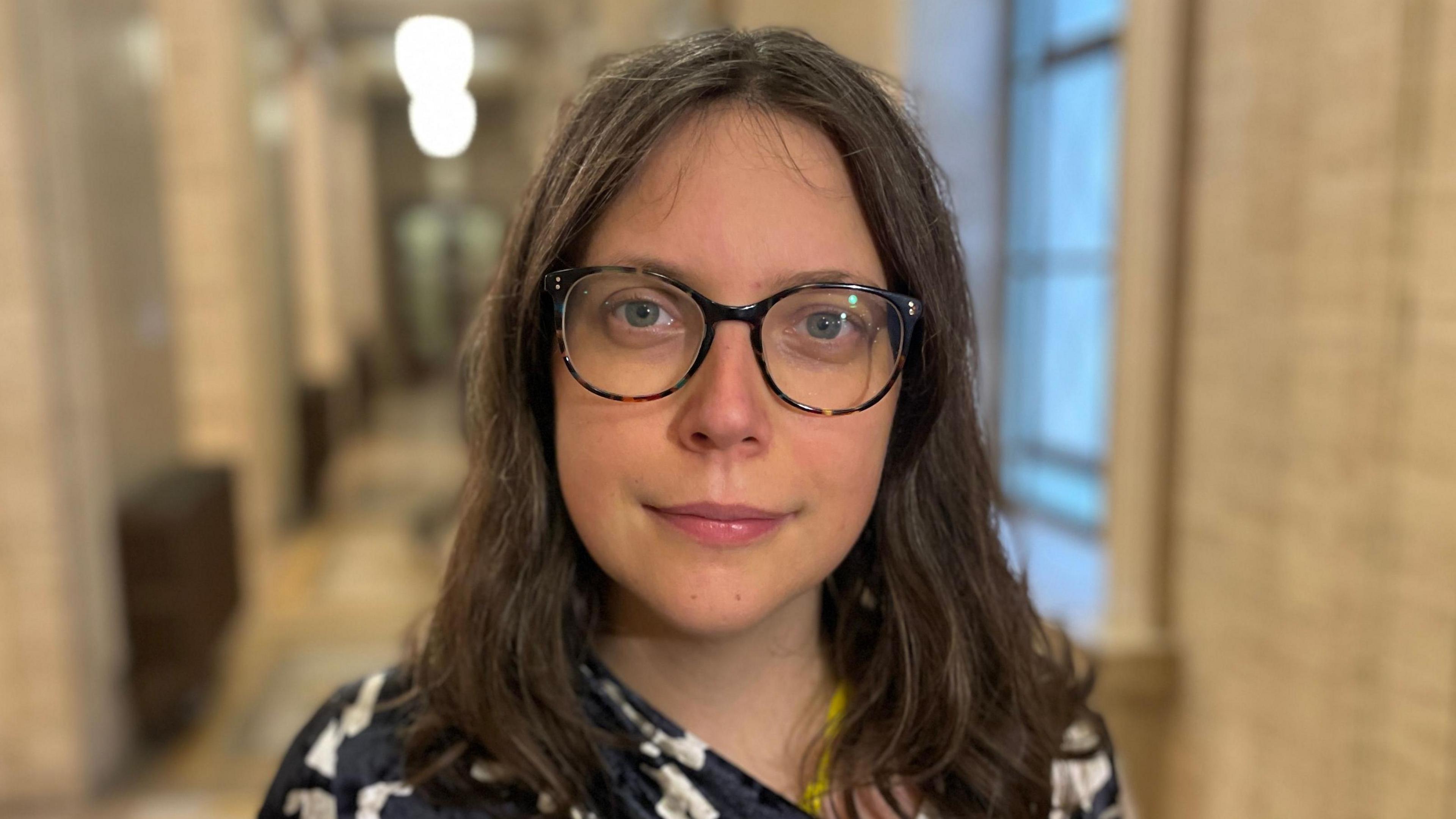 Dr. Viviane Gravey inside Stormont, she has long brown hair and is wearing dark framed glasses. Her top is black and white patterned. 