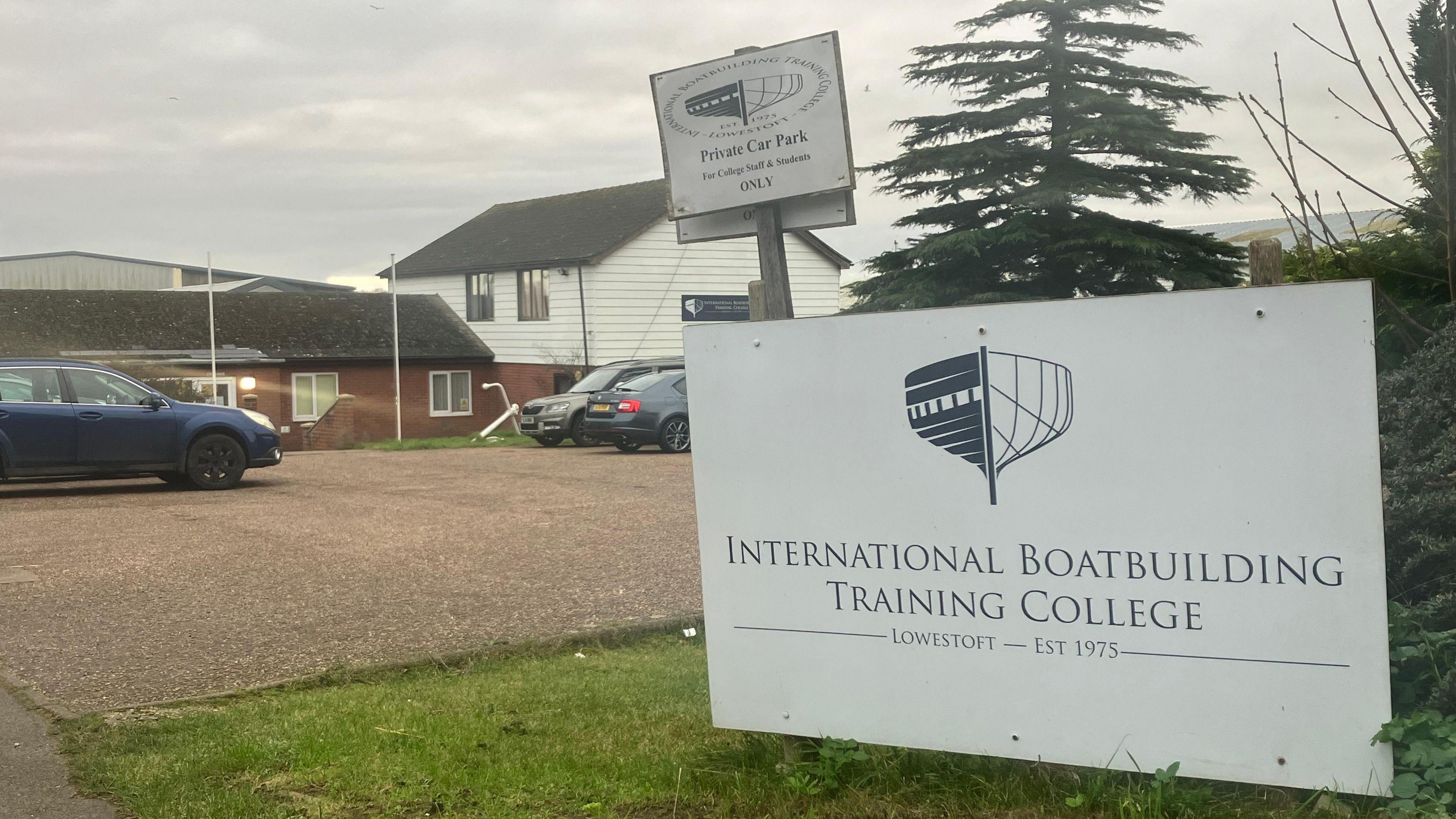 A large white sign reading International Boatbuilding Training College is situated on a grassy area next to a car park, with college buildings in the background 