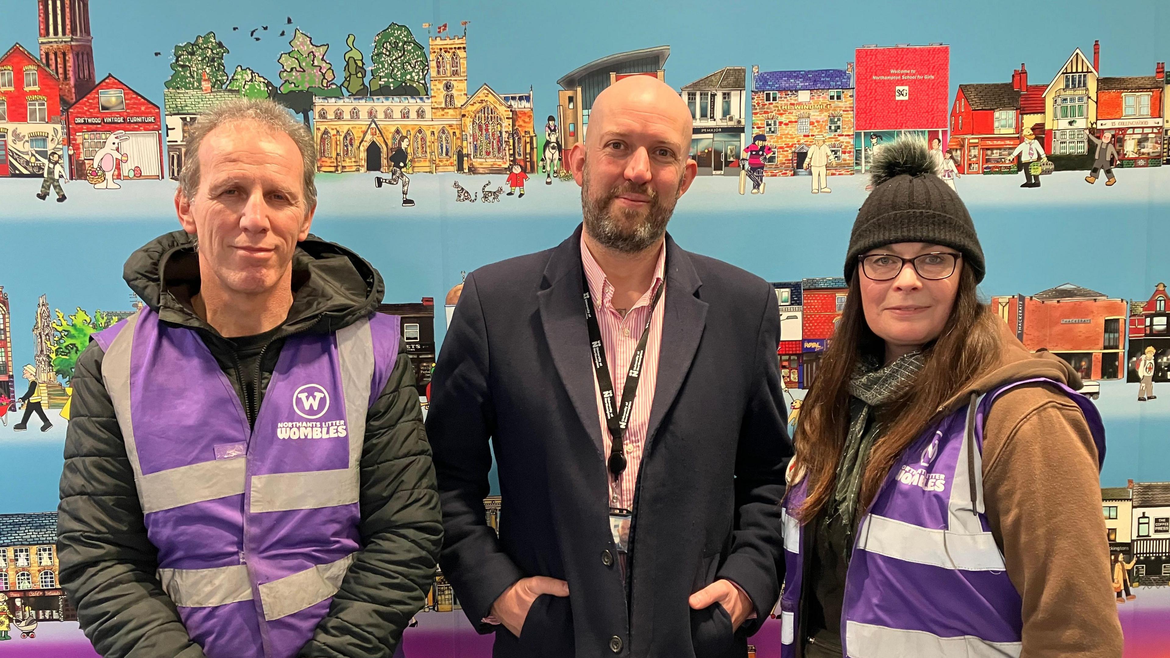 A group consisting of a man on left wearing a purple tabard labelled 'Northampton Litter Wombles' standing next to another man, who is wearing a black suit with a pink pinstriped shirt. A third person, a woman, is standing to the right with long brown hair and wearing the same purple tabard. 