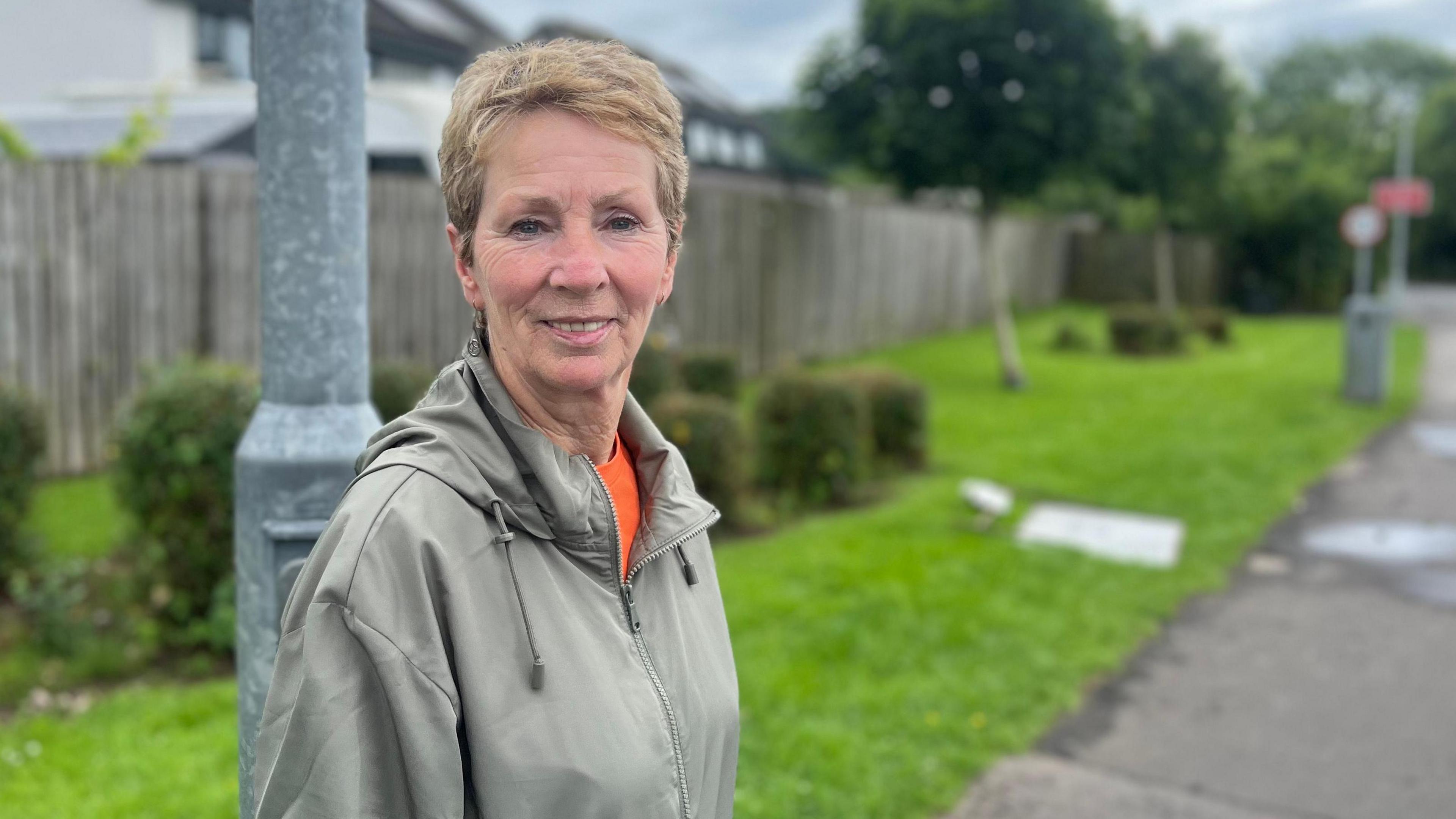 A woman with short reddish-blonde hair in a pale green anorak with an orange top underneath stands in front of a lamp post and pathway