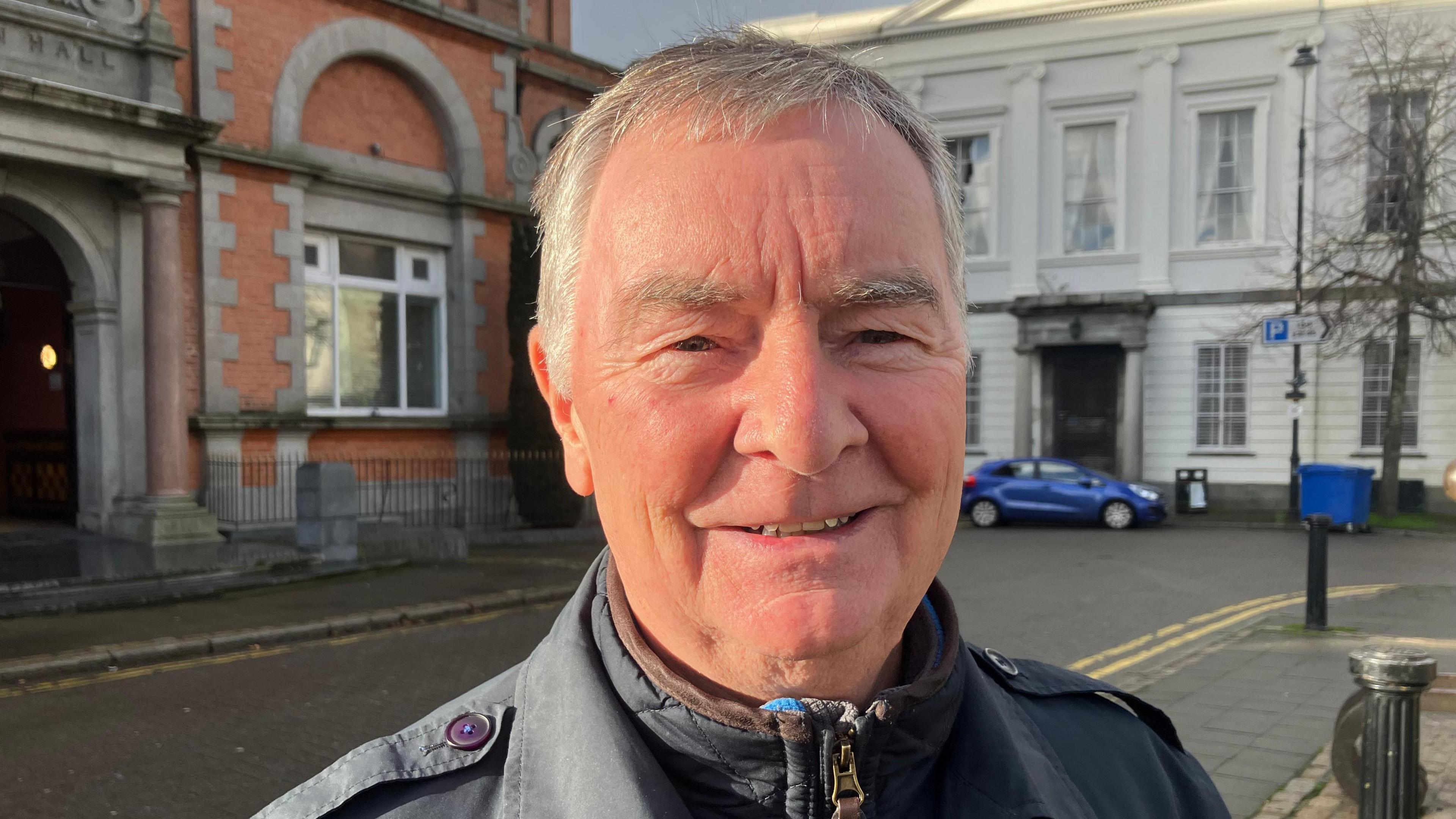 Sean Treanor has short grey hair and is pictured smiling.
He is stranding in Newry city centre while wearing a navy overcoat.
A large white building with a blue car is visible in the background.