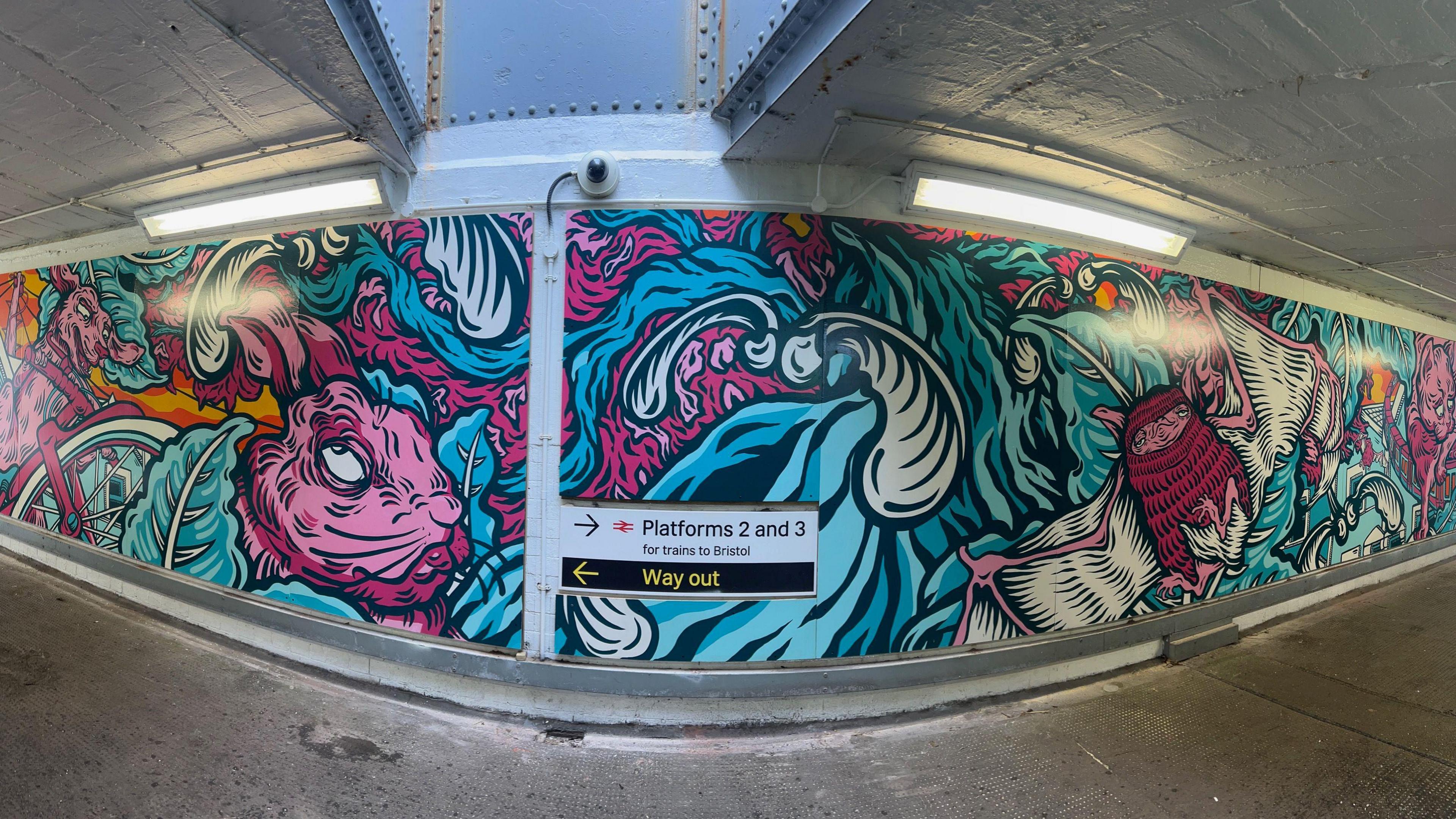 Image shows the Bedminster station underpass mural, in pink and blue colours. Images of rabbits and bats cover the wall, with a sign for platforms 2 and 3 in the centre of the picture. 