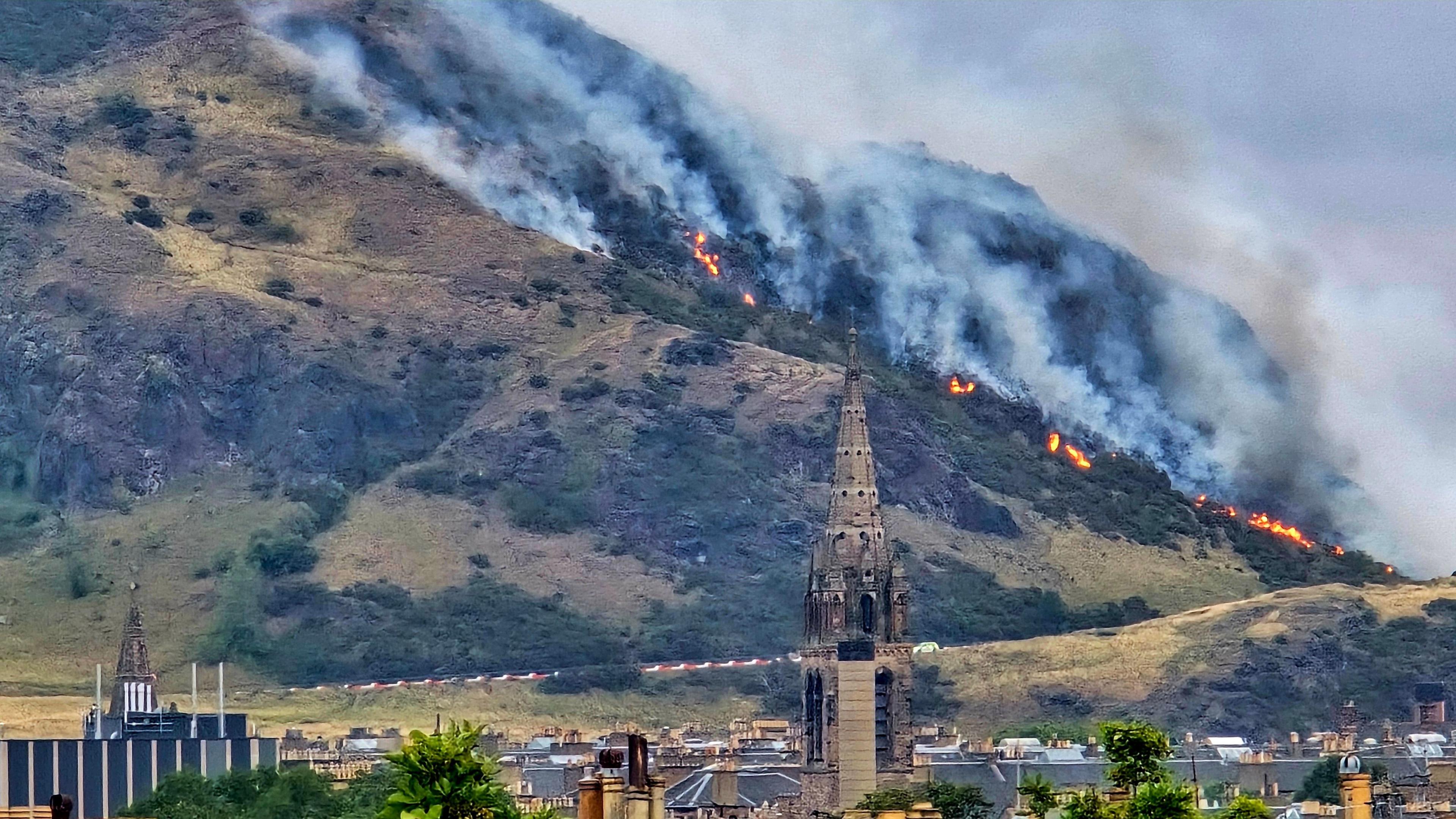 A fire burns on a grassy hill behind a church spire and other buildings in the foreground. The fire extends from the base of the hill further up the slope on the left, with thick grey smoke rising above it. There are large patches of orange flames.