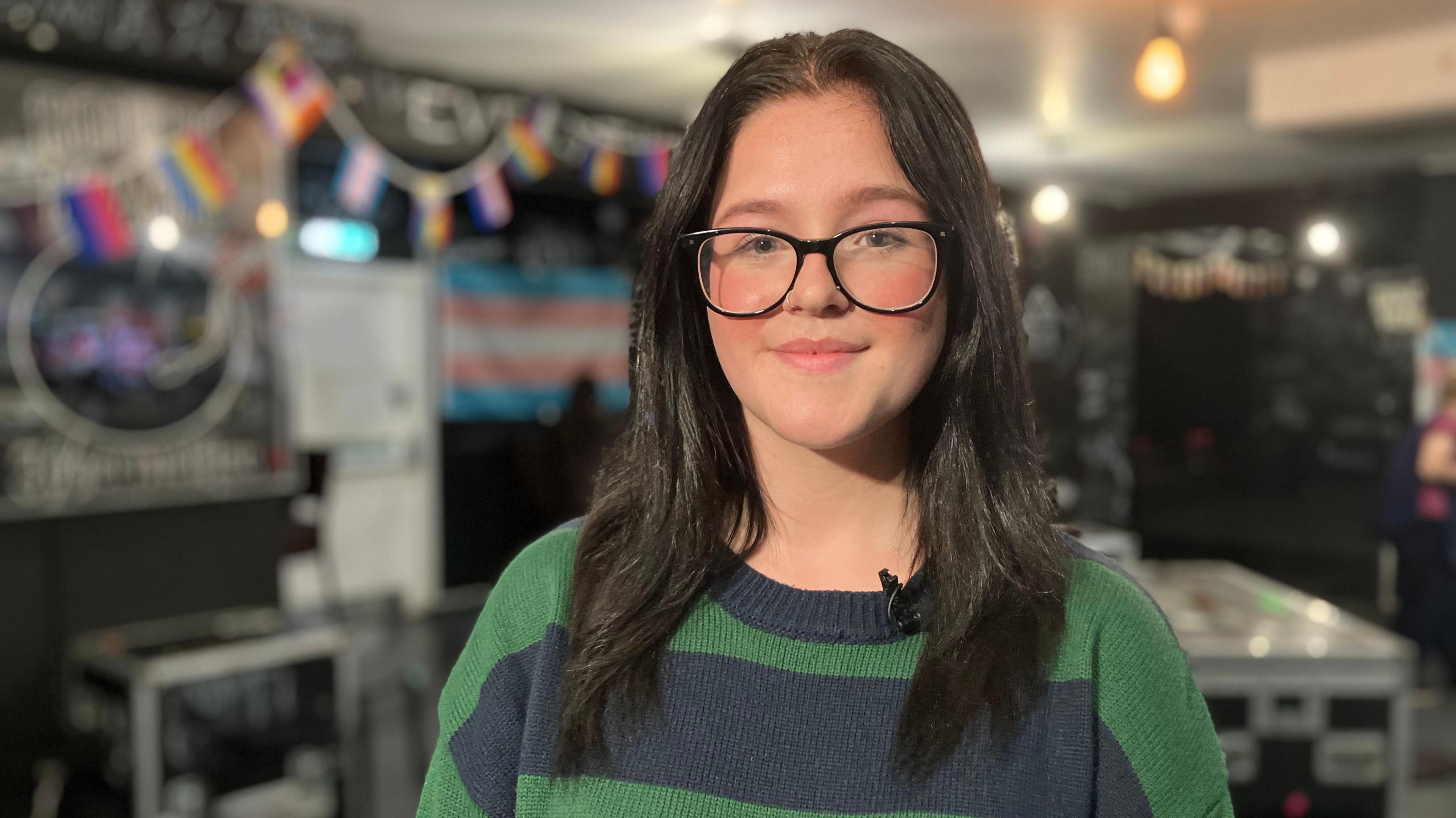 Hermione smiles and looks into the camera. She is wearing glasses with a green and blue jumper.