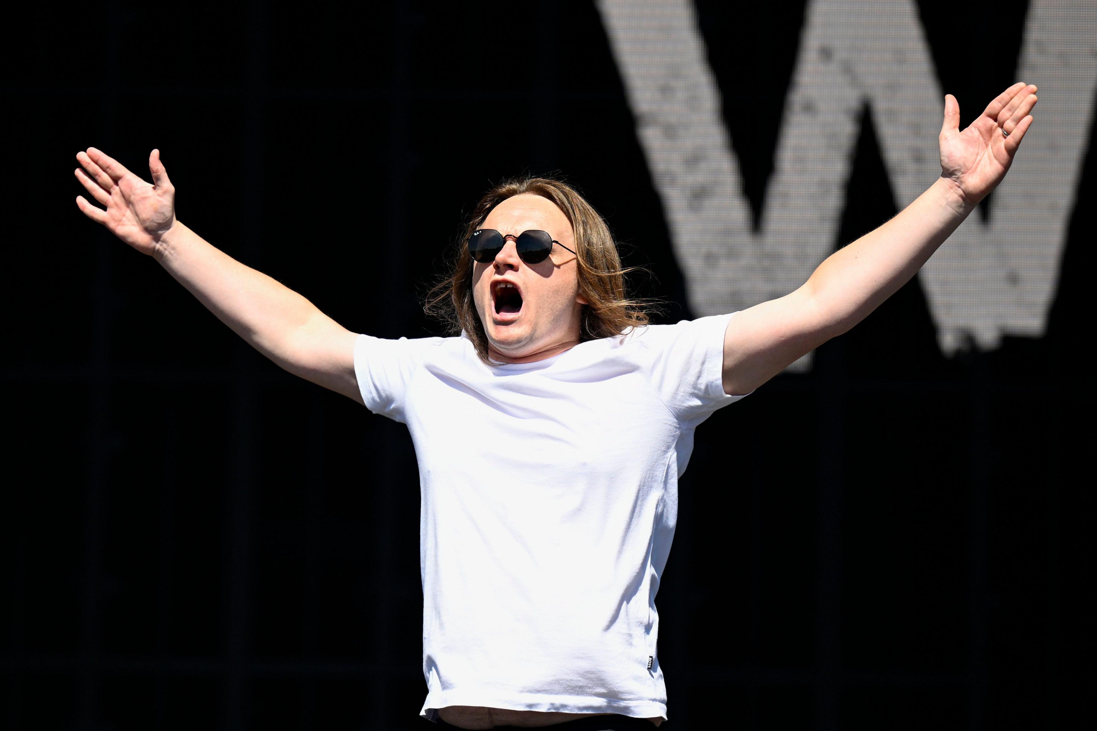 Jamie Webster is a young man in a white T-shirt with dark round sunglasses on his face. He is standing before a black backdrop with a big white W visible. He is holding his arms wide and high up.