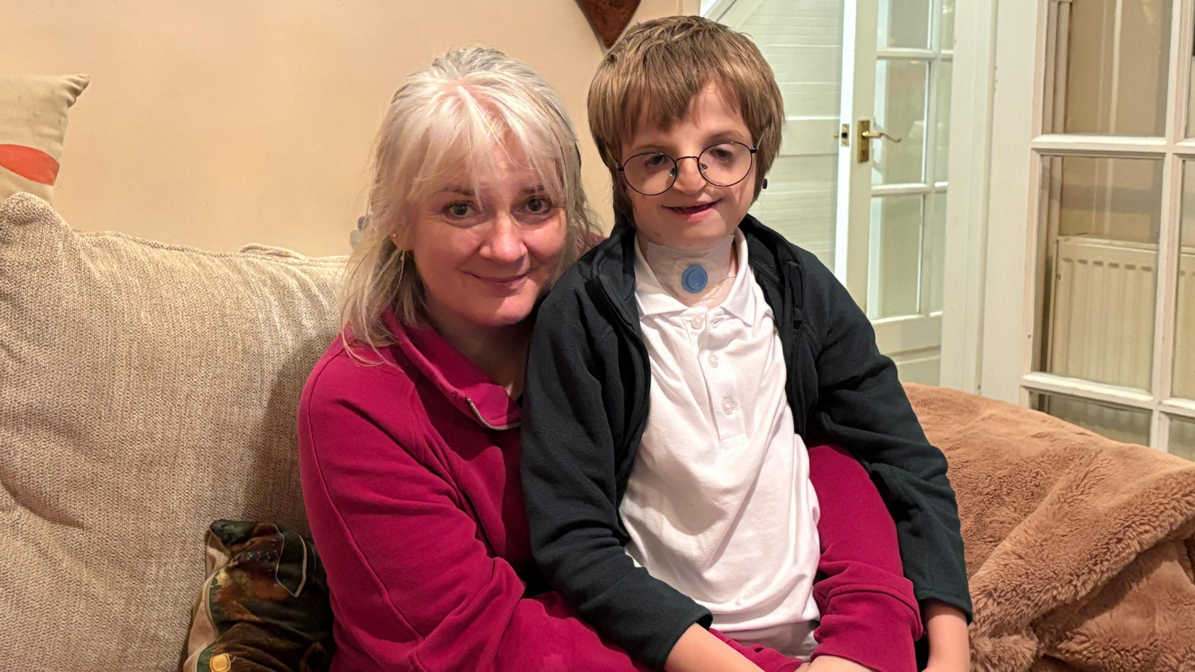 A boy with a genetic condition is wearing small black-framed glasses, a white polo-shirt and a black fleece. He is smiling at the camera while sitting on the lap of his mum who is wearing a pink jacket. She has grey hair and is also smiling. They are both sitting in a lounge on a beige sofa which has a light-brown fluffy blanket.