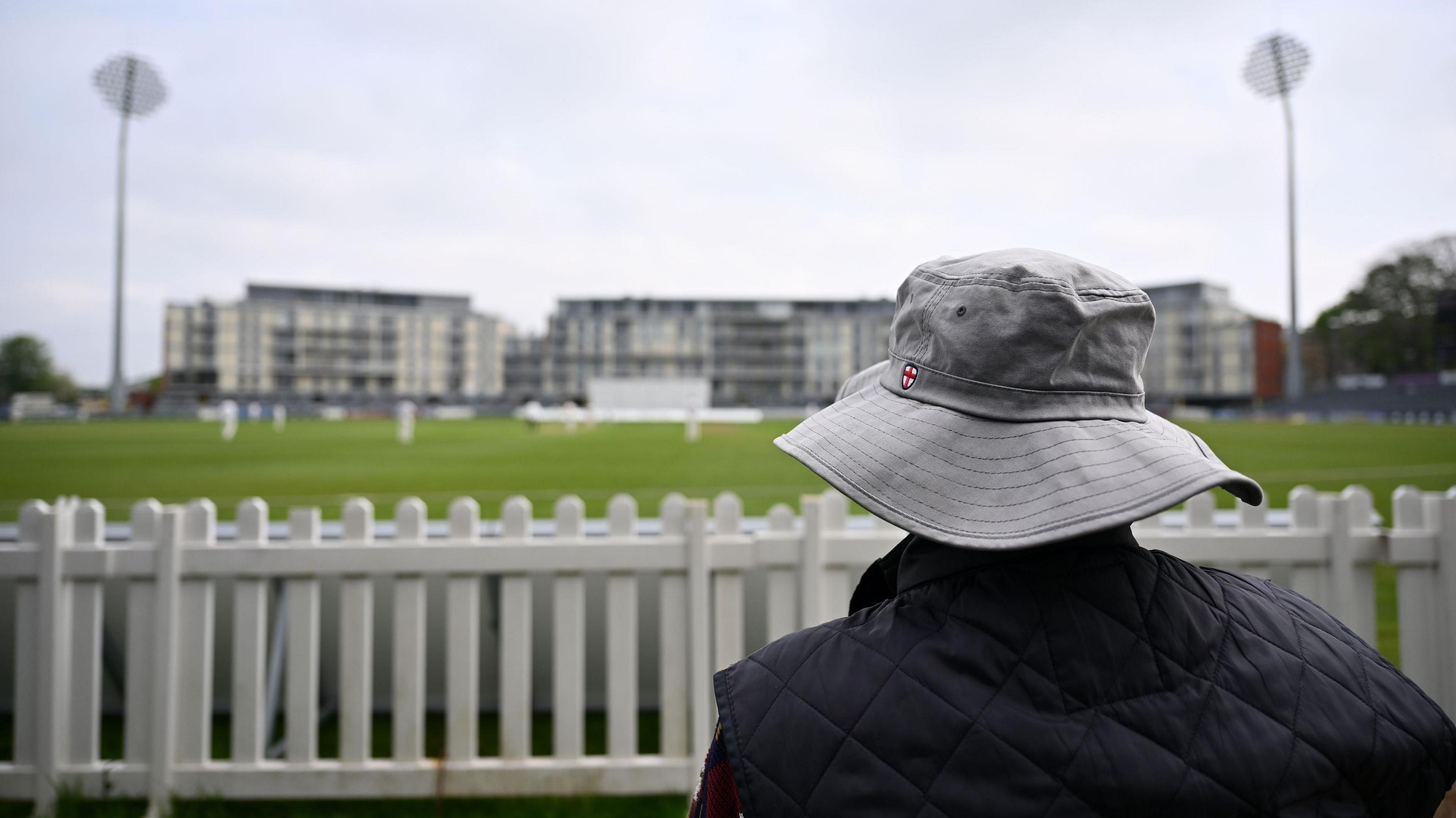 A fan wearing a bucket-style hat watches Gloucestershire v Leicestershire at the County Ground in Bristol. The fan is pictured from behind and the players and floodlights are visible, but not in focus, in the background