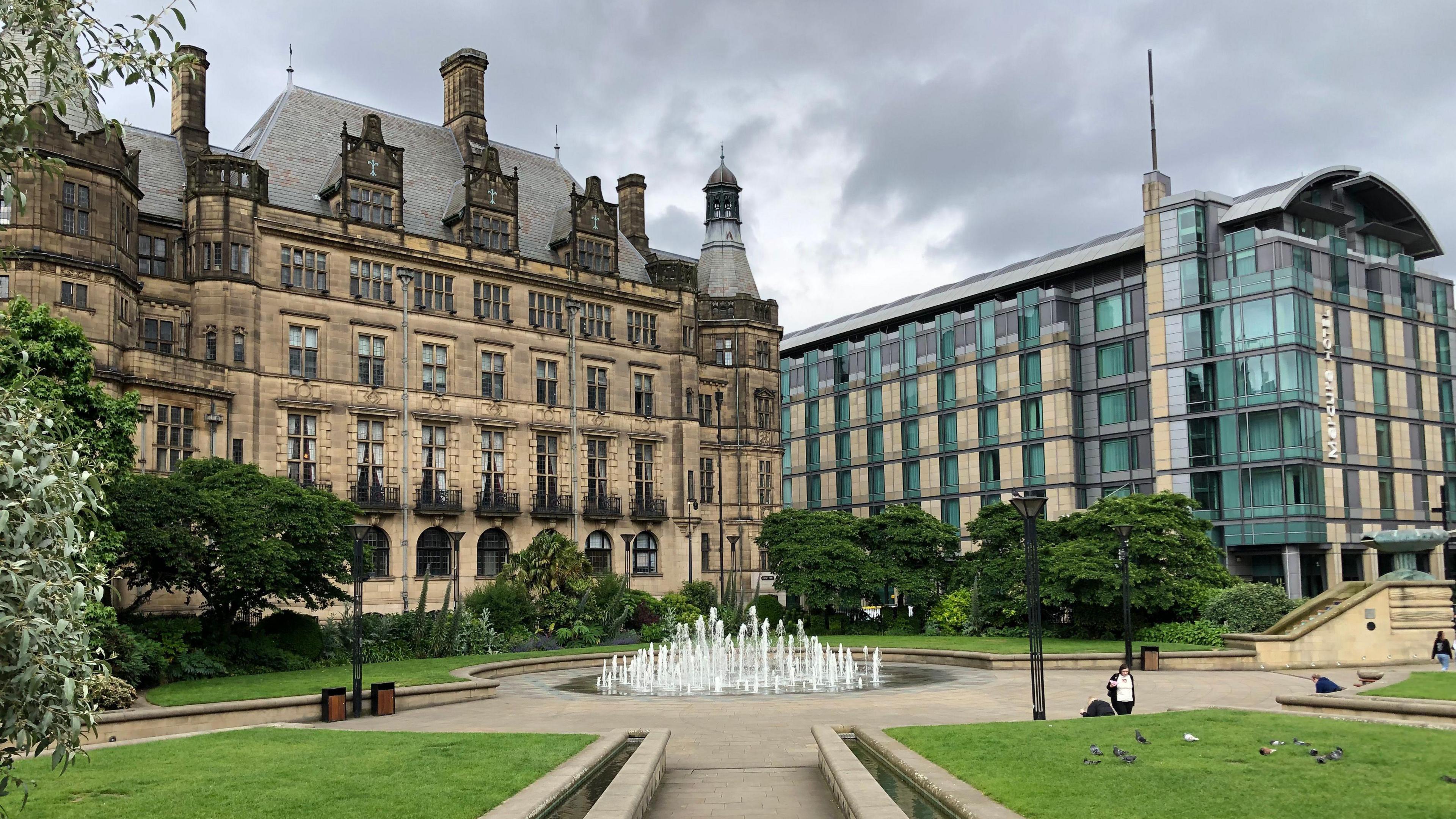 An image of Sheffield town hall and neighbouring Mercure hotel in Sheffield.  The Town hall is a large ornate Victorian stone building.  The hotel is a modern block of cream coloured stone and light blue glass.  In front of the buildings is a fountain and grass areas, which make up the Peace Gardens