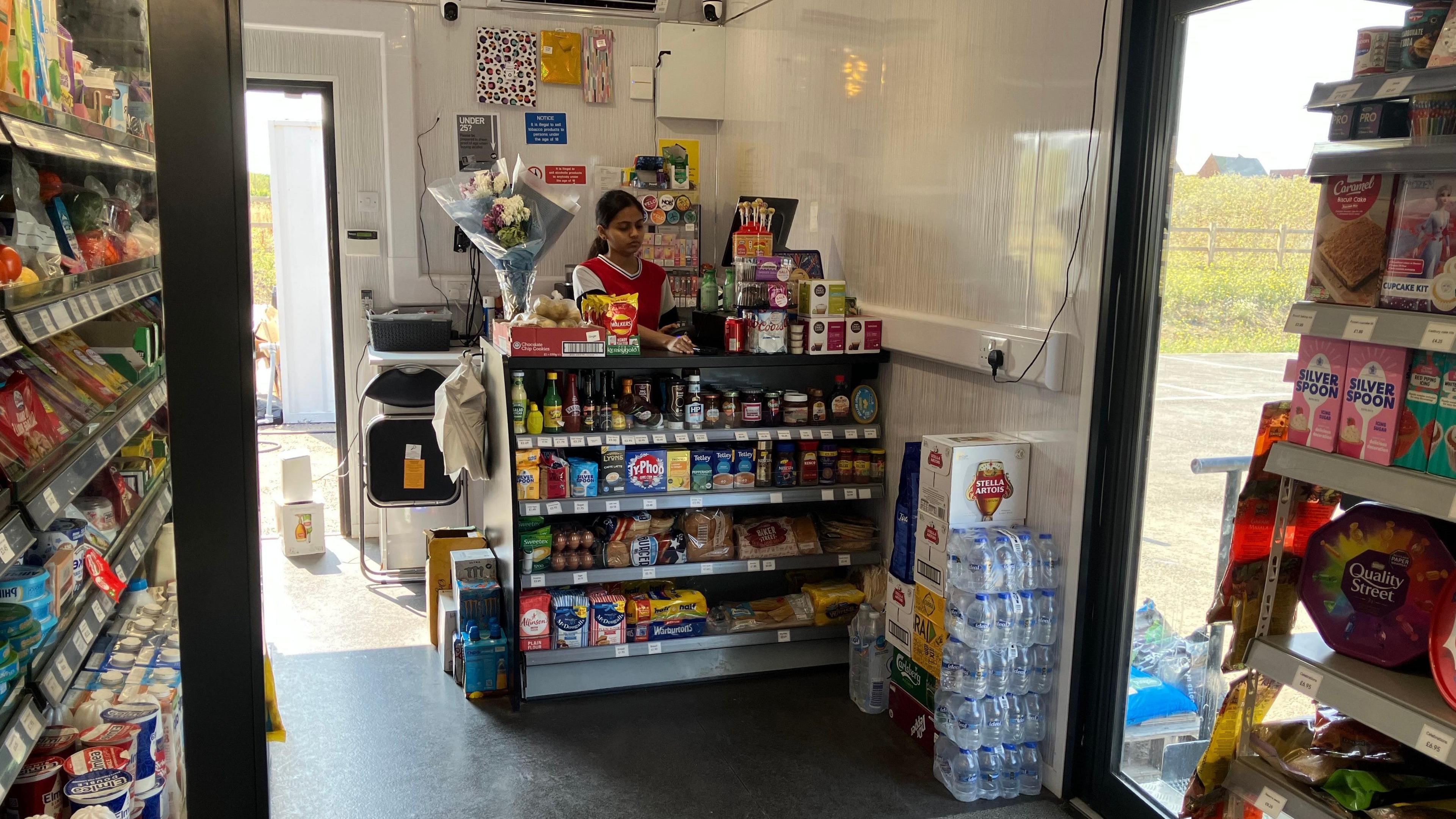 A small narrow shop. To the left of the picture is a fridge where items like sausage rolls and soft cheese can be seen. To the right shelves display items including sugar and boxes of chocolate. In the middle of the picture is a counter with groceries on shelves underneath. A worker is standing at the counter in a red apron. The counter has other items for sale including a bunch of flowers. Two doors can be seen, one at the back of the picture and one to the right. There are various posters on a white wall at the back of the picture.
