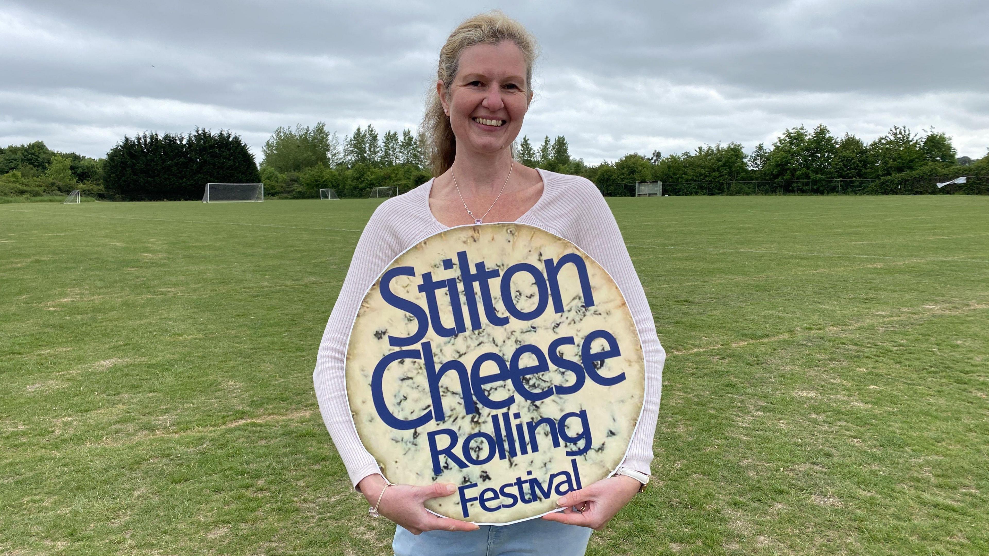 Sara wearing a white long-sleeved T-shirt and blue jeans holding a round block of yellow cheese with Stilton Cheese Rolling Festival written on it in purple