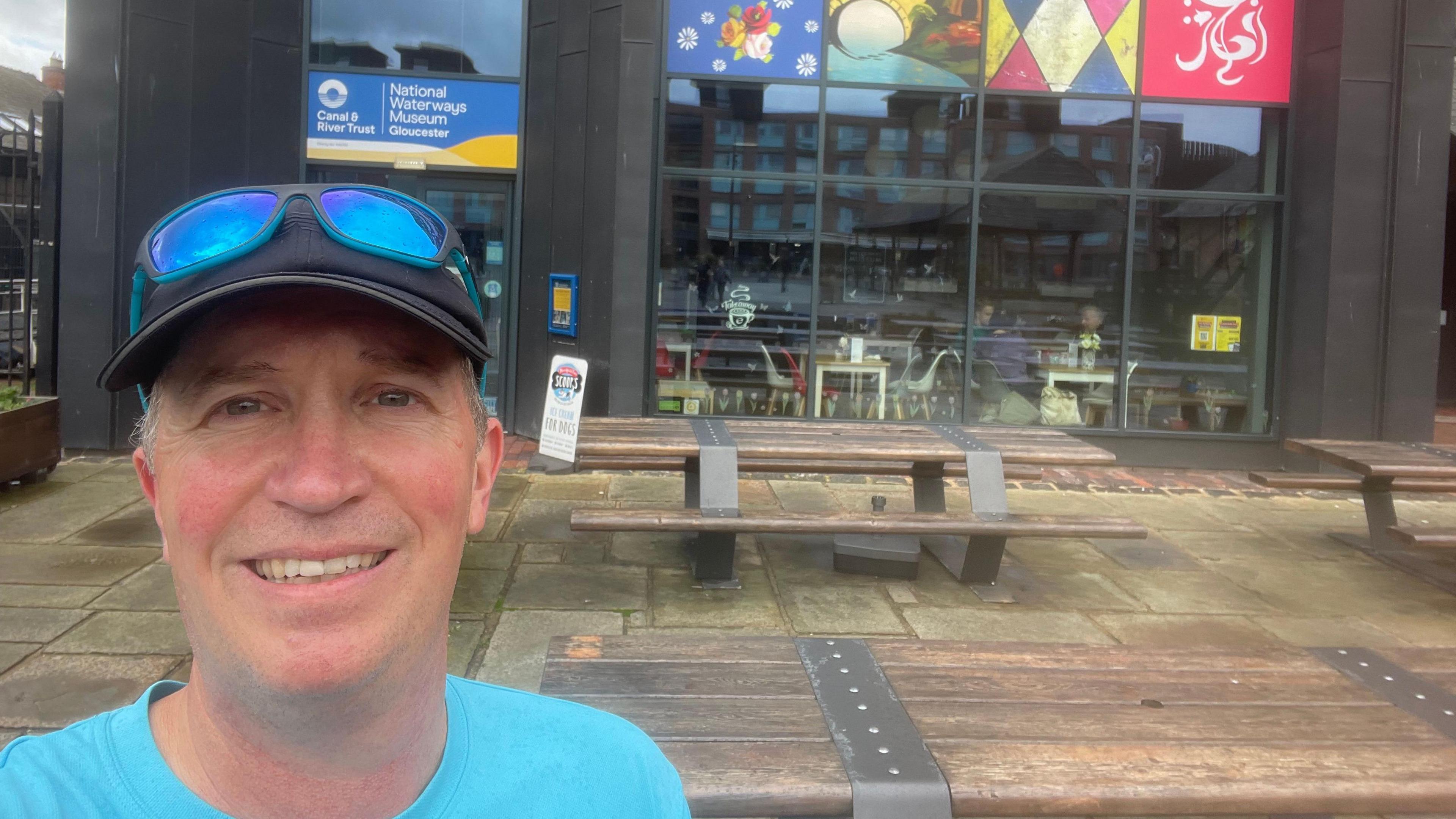 Matt Williams smiles at the camera in the foreground, wearing a blue t-shirt and a black cap. Behind him is the front of the National Waterways Museum.