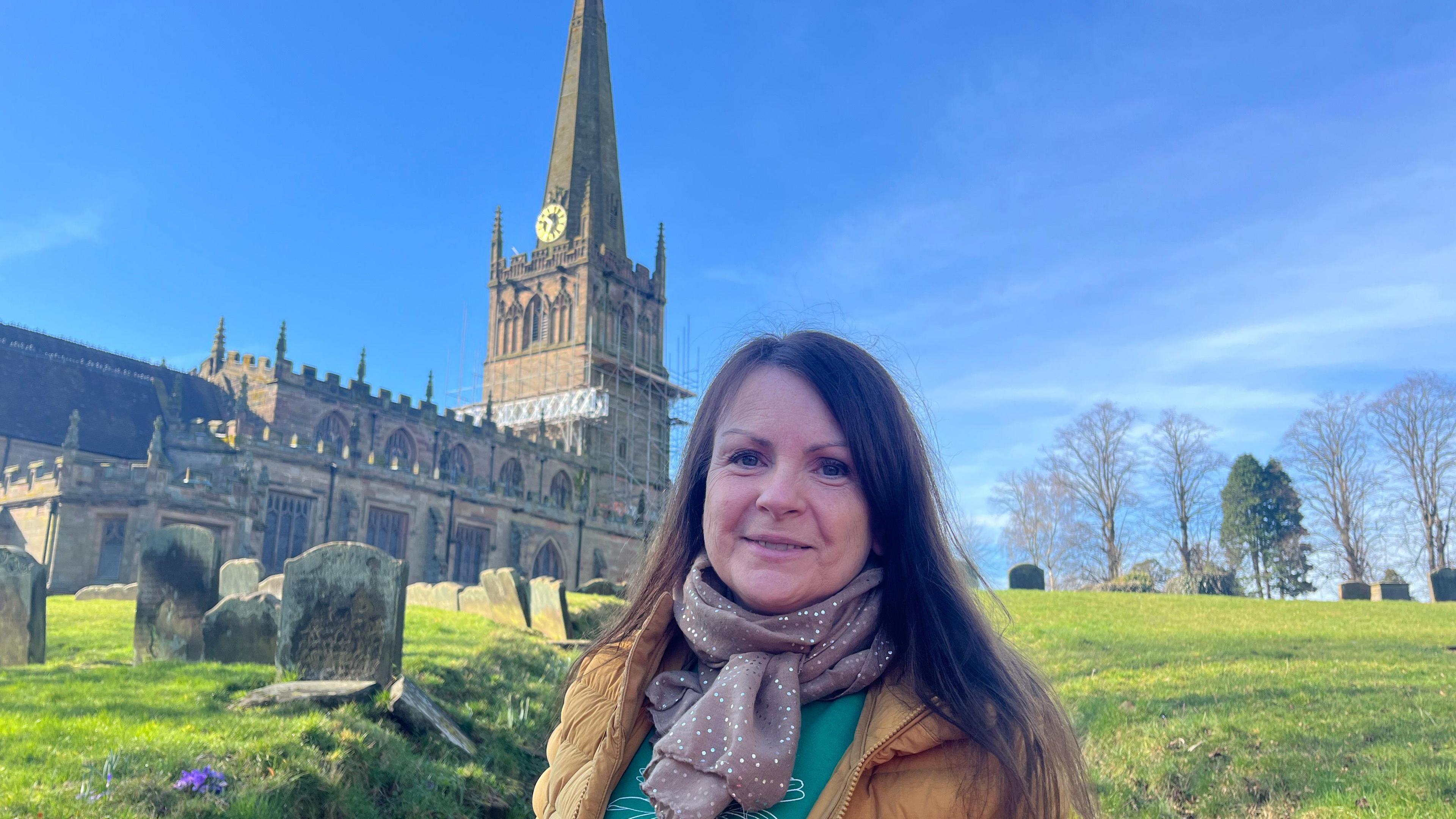 A brunette woman is smiling in front of a church with scaffolding on its spire. She wears a green top with a flower, a yellow puffer jacket, and a brown sparkly scarf. The sun is shining and the sky is clear blue.