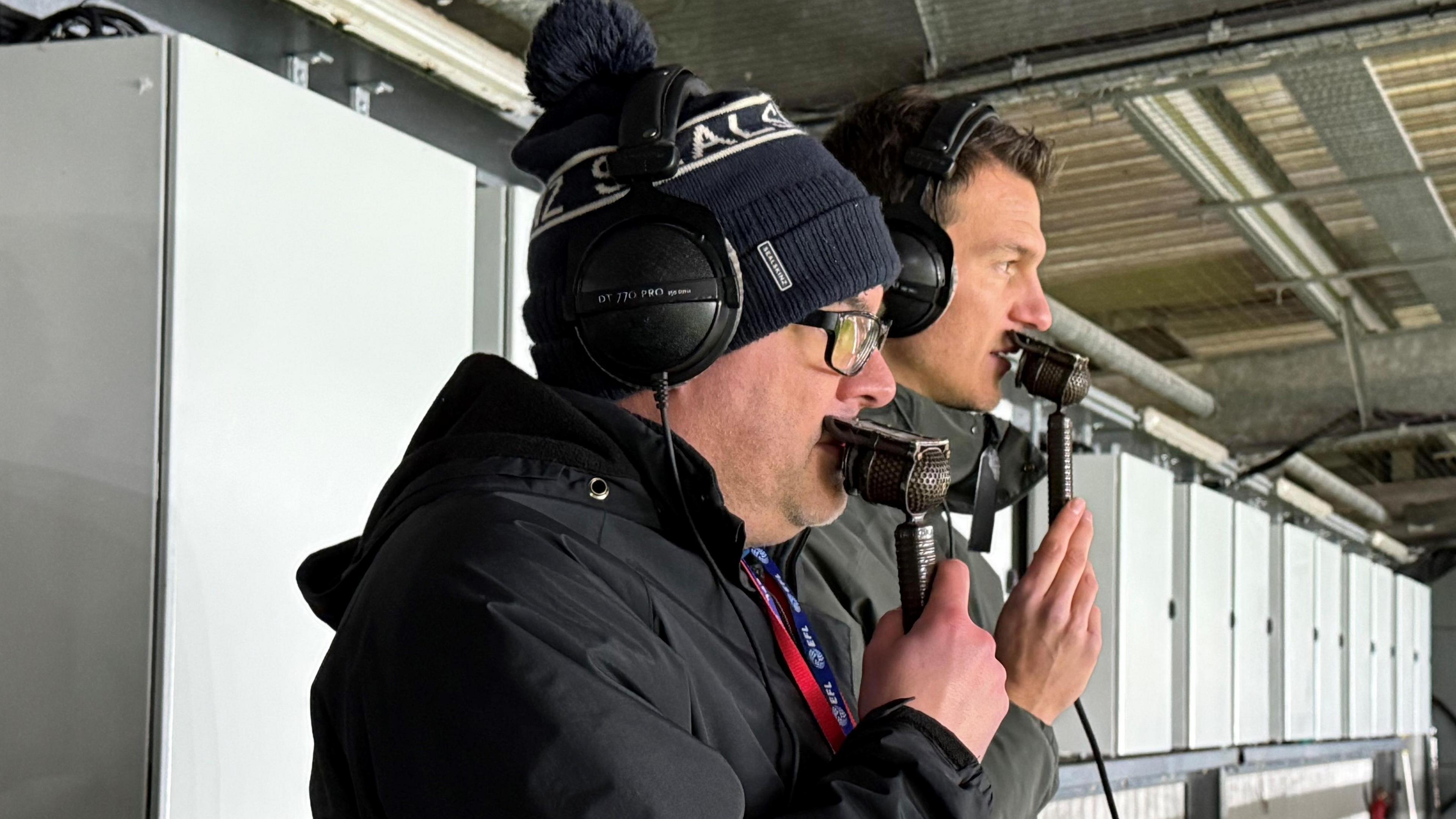 A man with glasses and a woolly hat holding a microphone to his mouth while standing on a gantry. Another man standing just behind him is also speaking into a microphone.