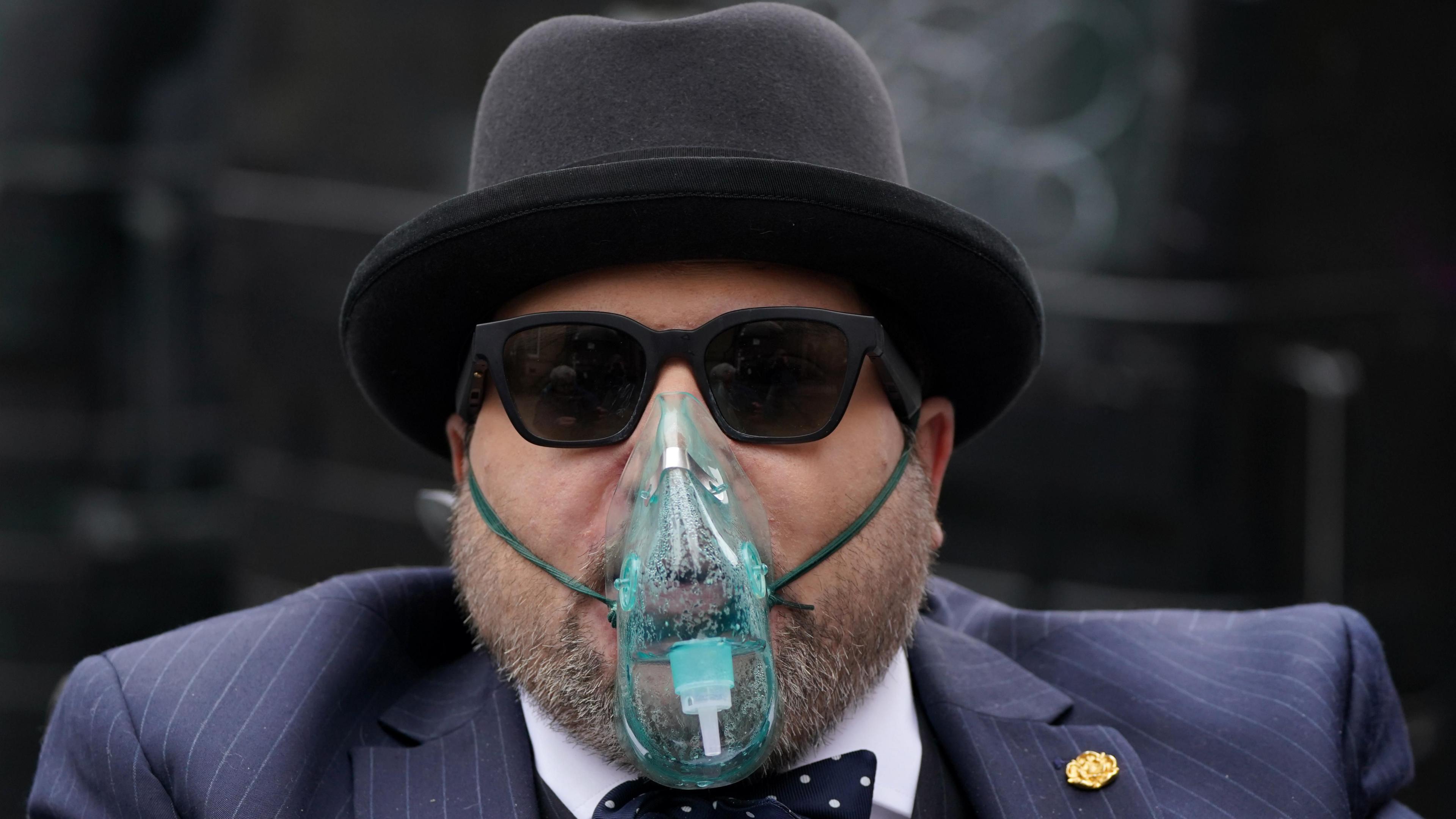 Nicholas Rossi wearing a pinstripe suit, black sunglasses, black hat and an oxygen mask. He has a grey beard.