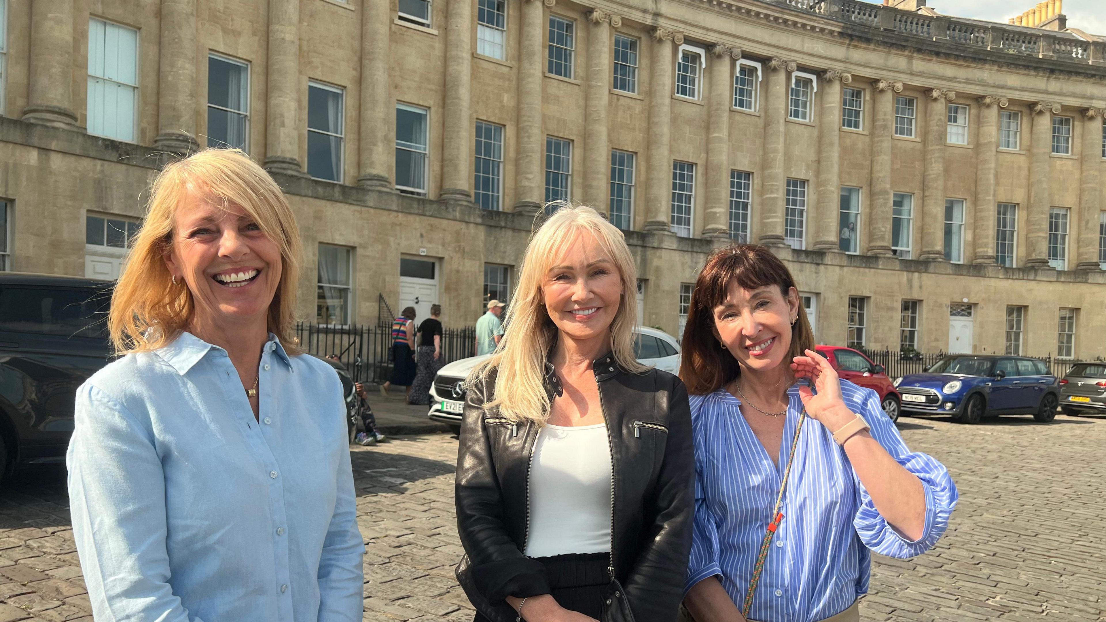 Three women stand outside their houses in the Royal Crescent in Bath smiling at the camera