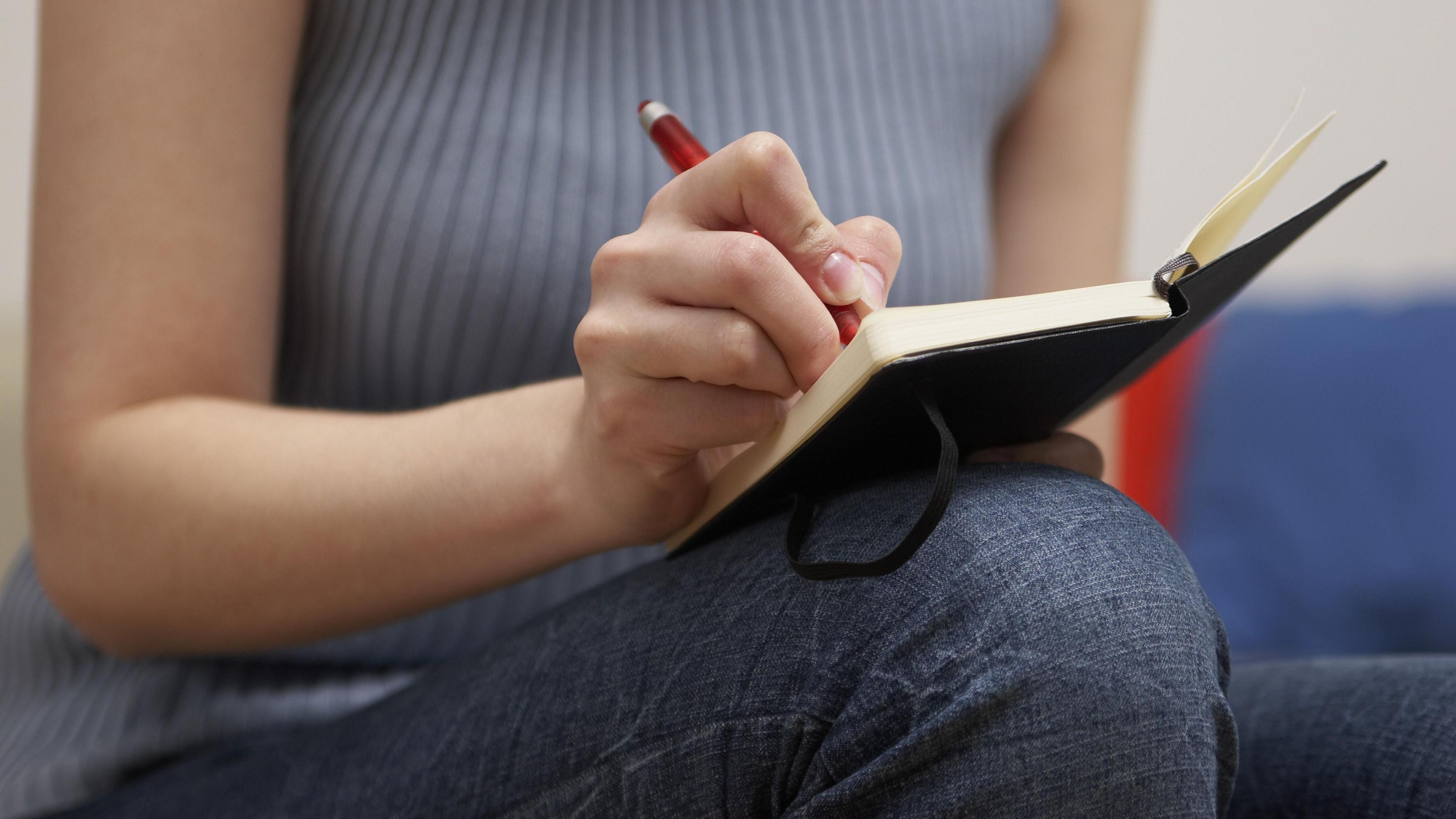 A close up of a woman's hand holding a pen as she writes in a black hard back notebook while sitting on a sofa.