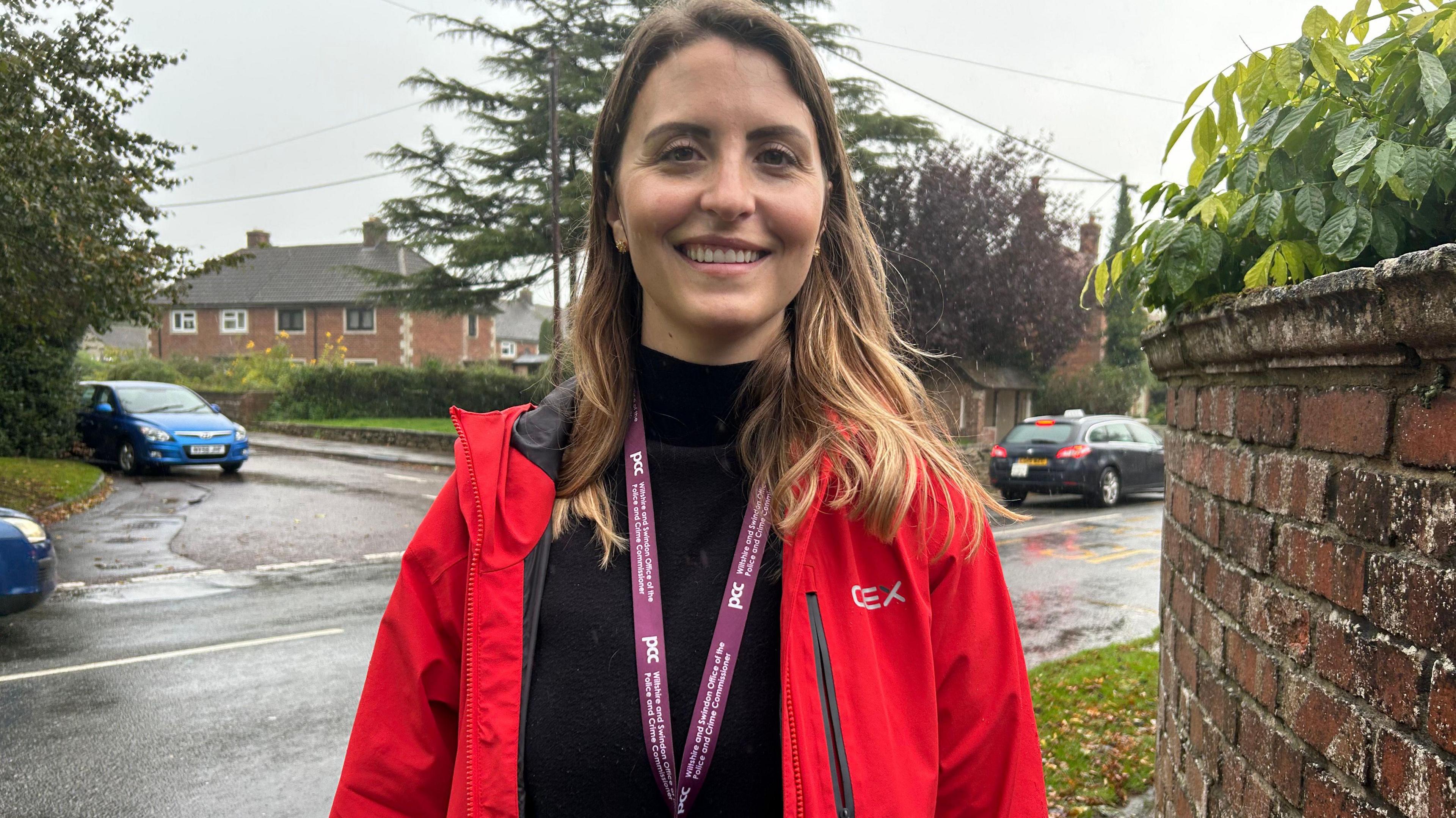 Natasha Gell has medium-length brown hair and is wearing a black high-neck top underneath a red jacket