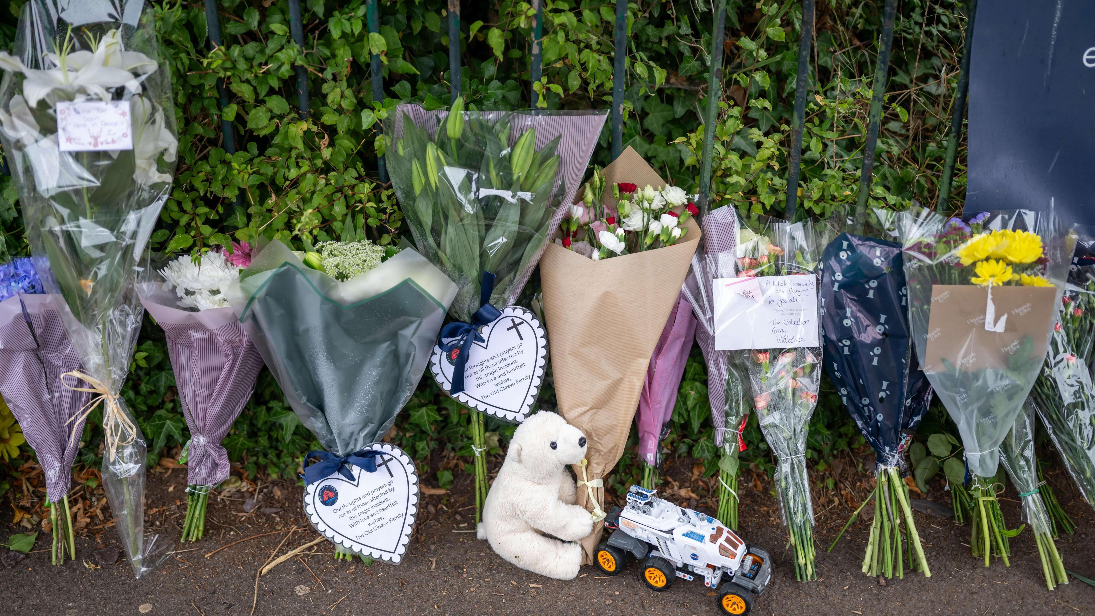 Several bouquets of flowers laid in tribute to a boy who died. Many of them include notes and cards. There is also a teddy and a toy car on the ground.