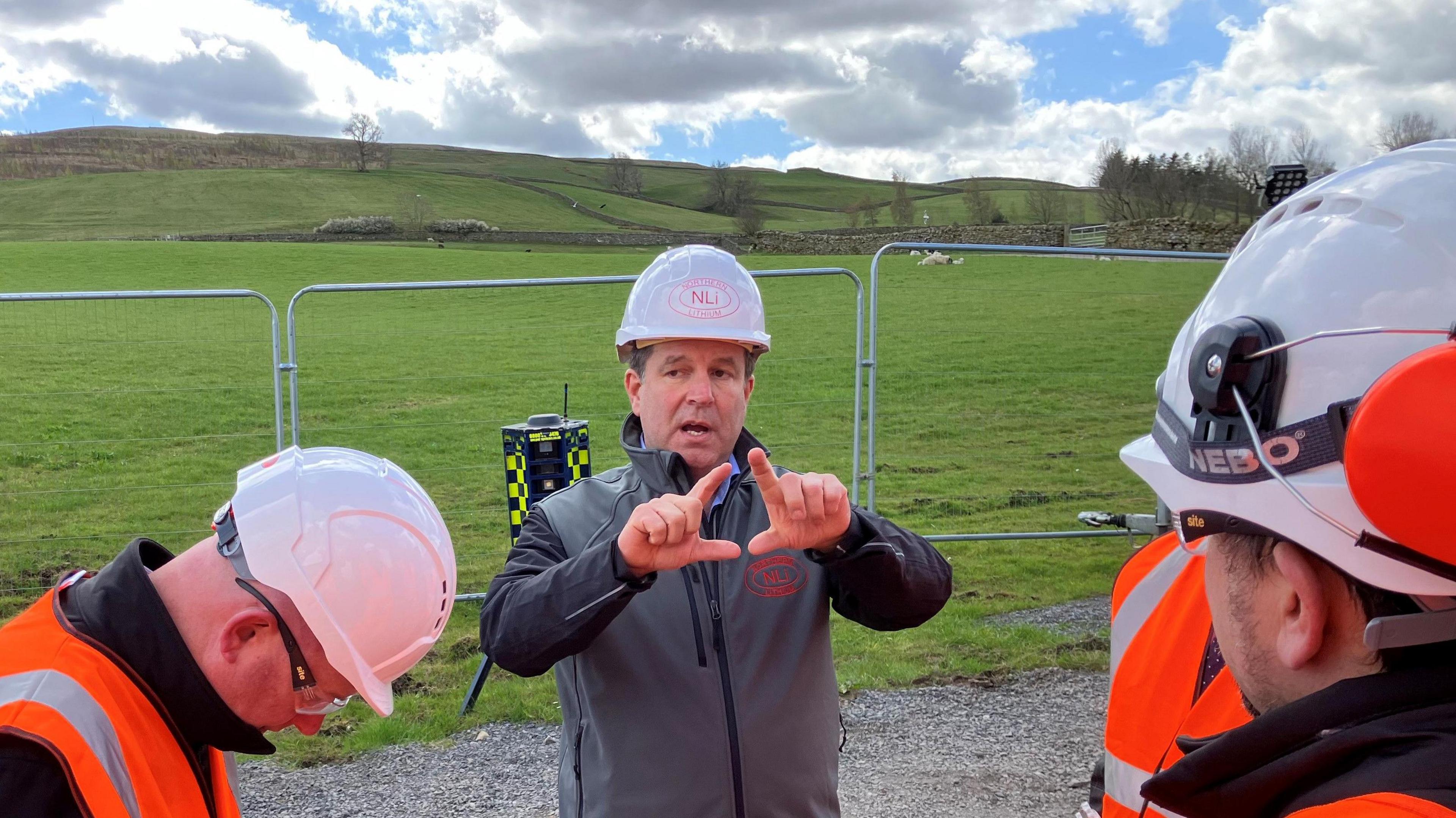 Nick Pople talking to workers and demonstrating with his hands at the site of the test plant. He is wearing a grey sweatshirt and white helmet. Workers in high visibility protective orange clothing have surrounded him. There is a large area of grassland behind him with several sheep resting nearby.