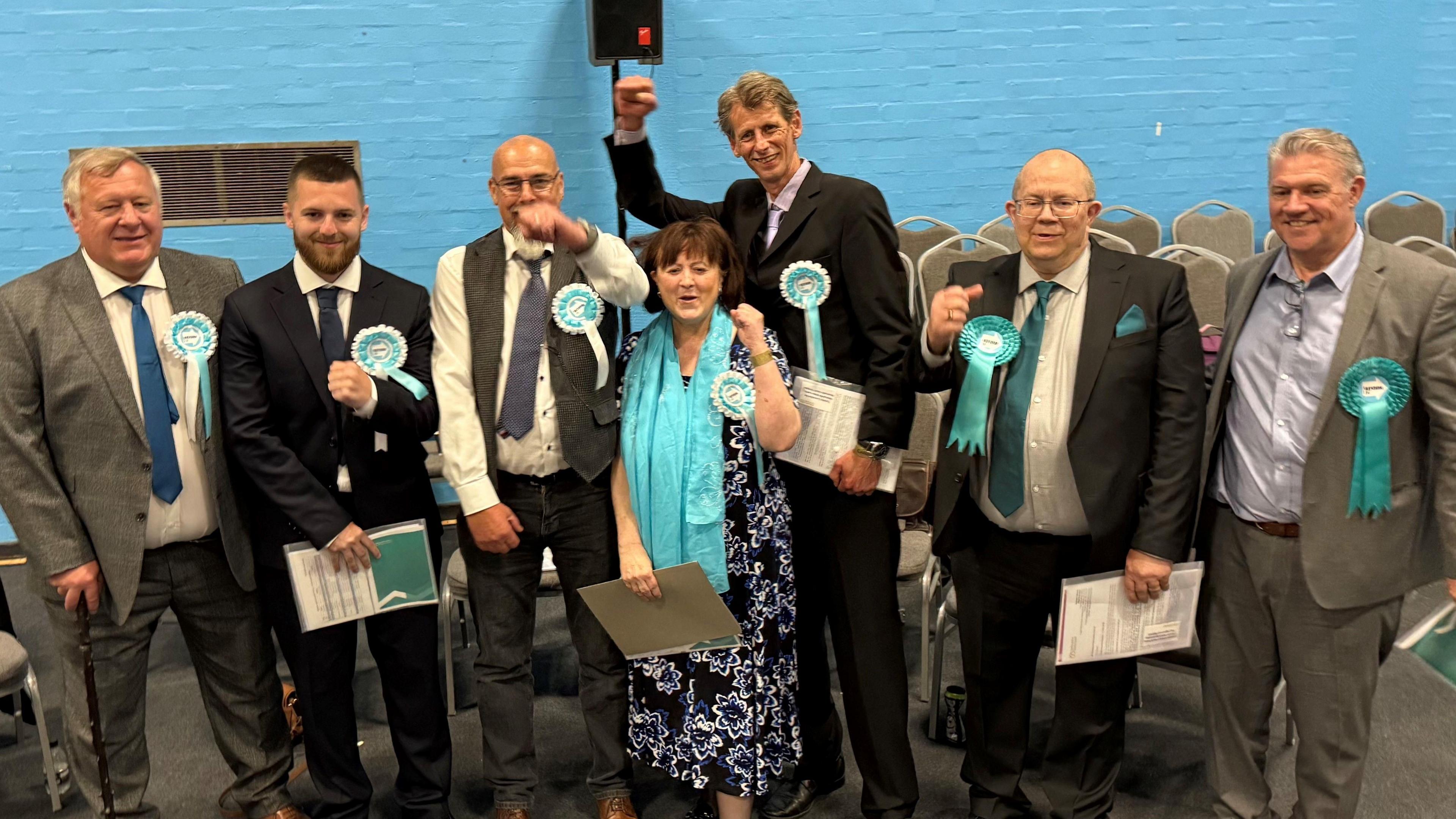 Seven Reform UK politicians are seen smiling and celebrating. They are wearing light blue rosettes and are stood against a blue wall.