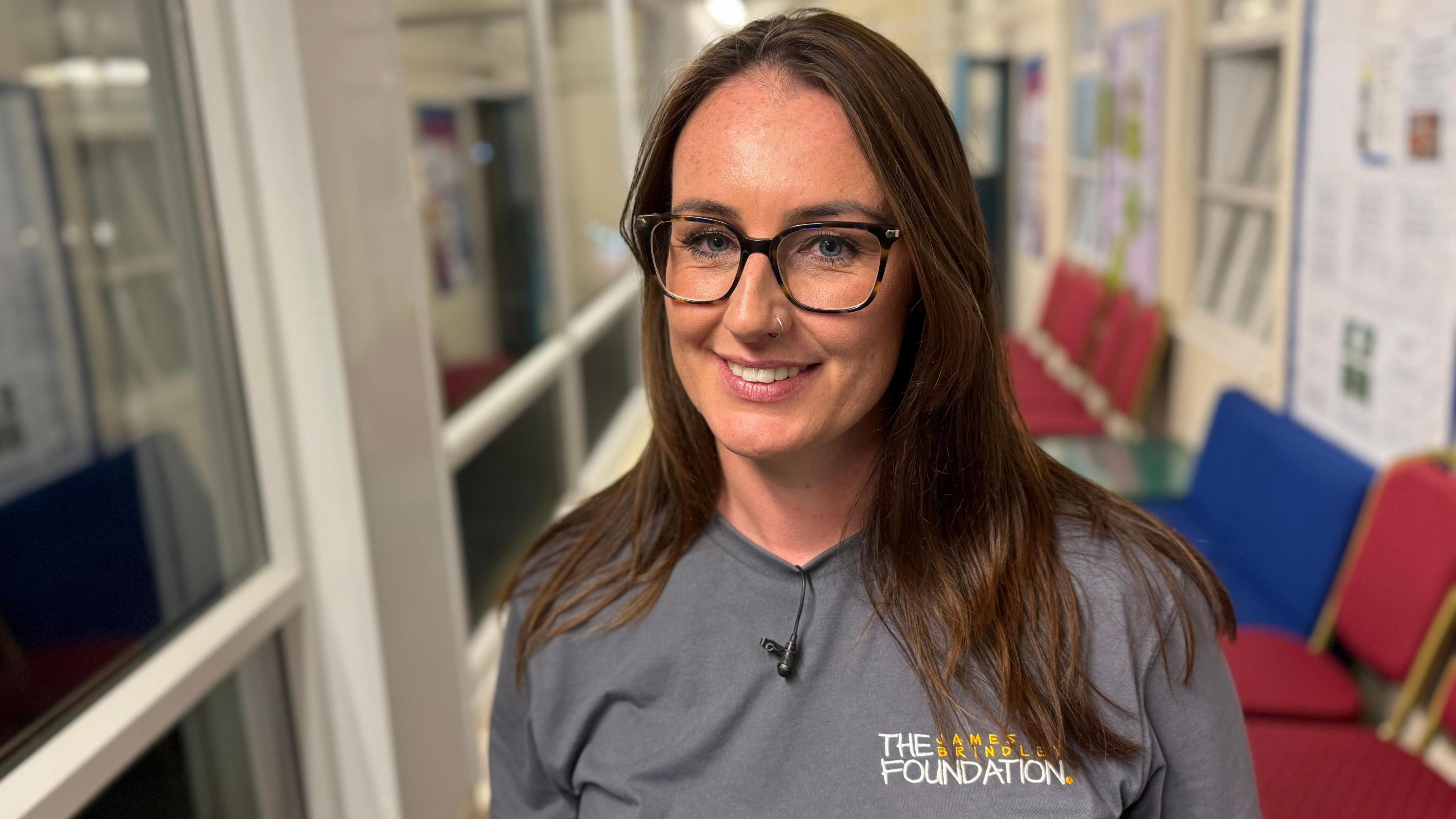 Nicola Neville smiles at the camera. She wears glasses and a grey T-shirt which says the James Brindley Foundation on it. 