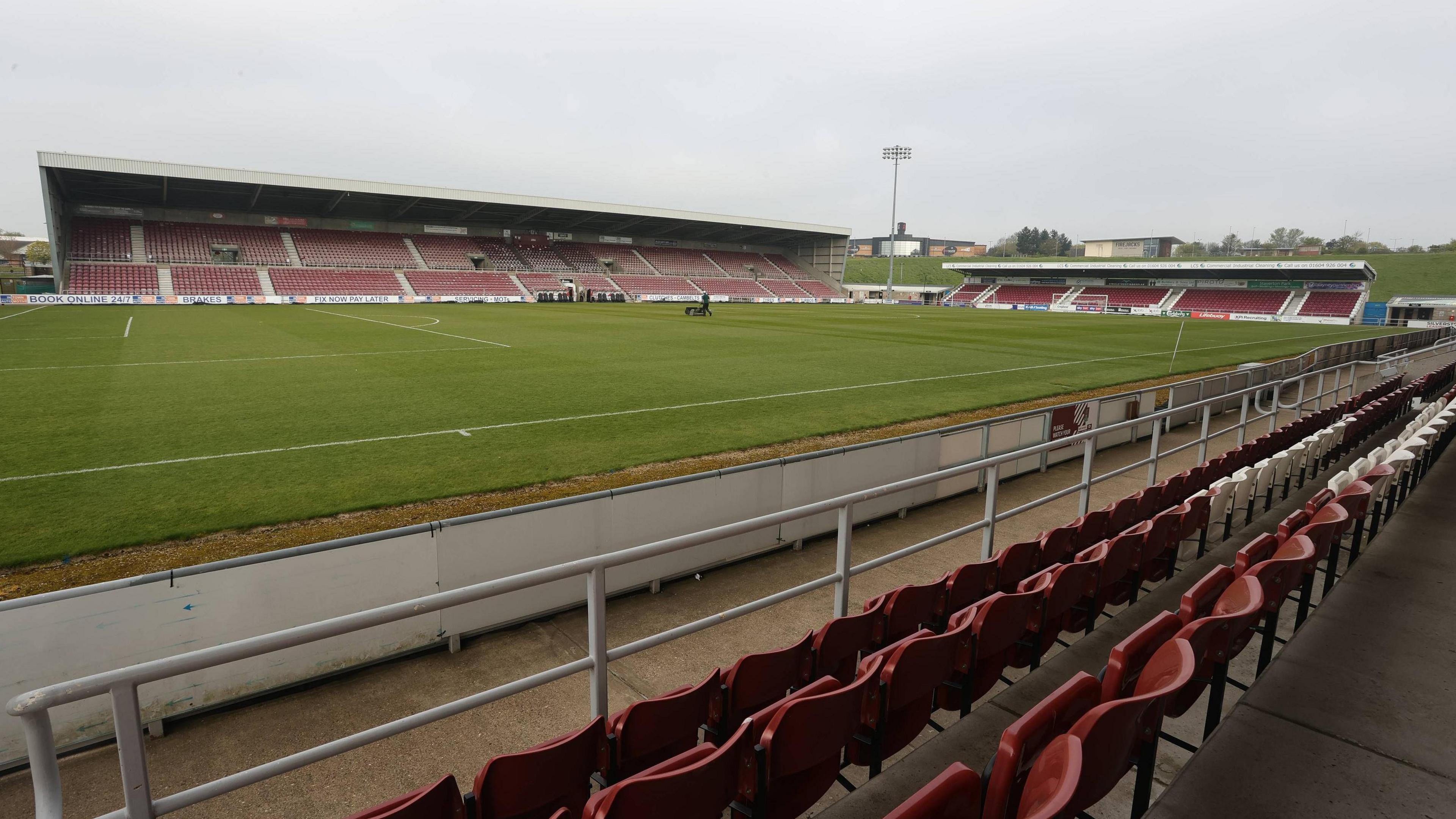 Sixfields Stadium as viewed from within the East Stand, looking towards the West Stand and North Stand. The stadium is empty, but for a lone groundsman mowing the pitch on an overcast day.