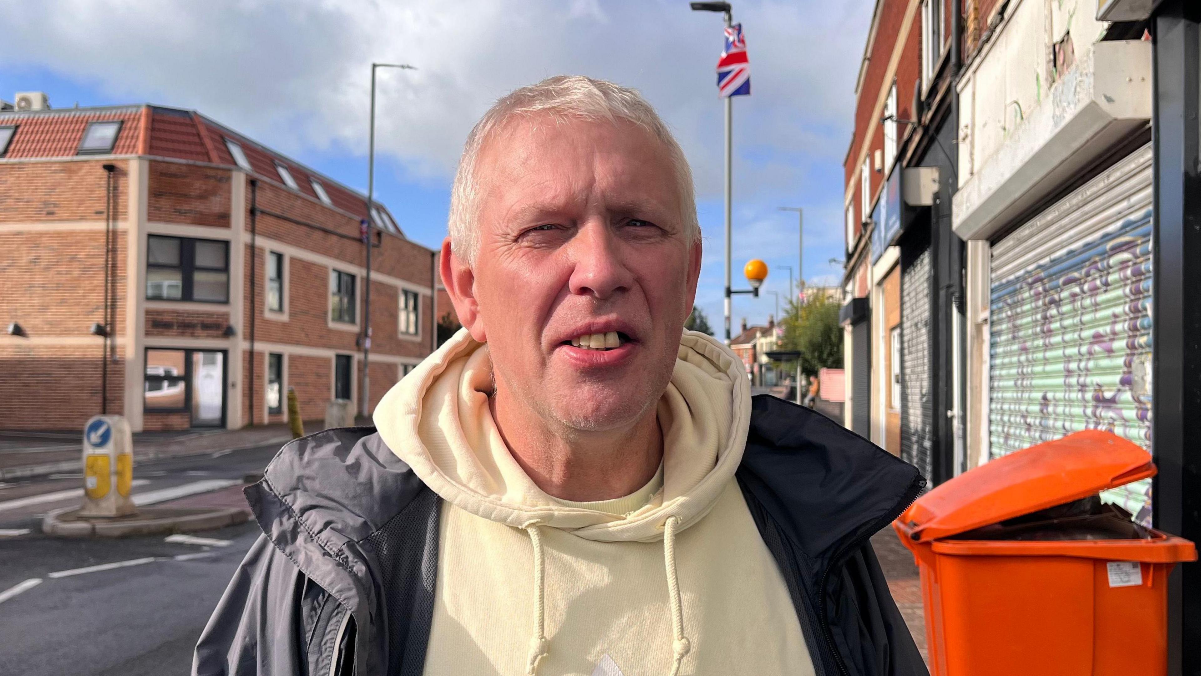 A man stands with a yellow hoodie and black jacket, behind them there is a road and next to the road a lamp post where a union flag hangs, blowing in the wind.