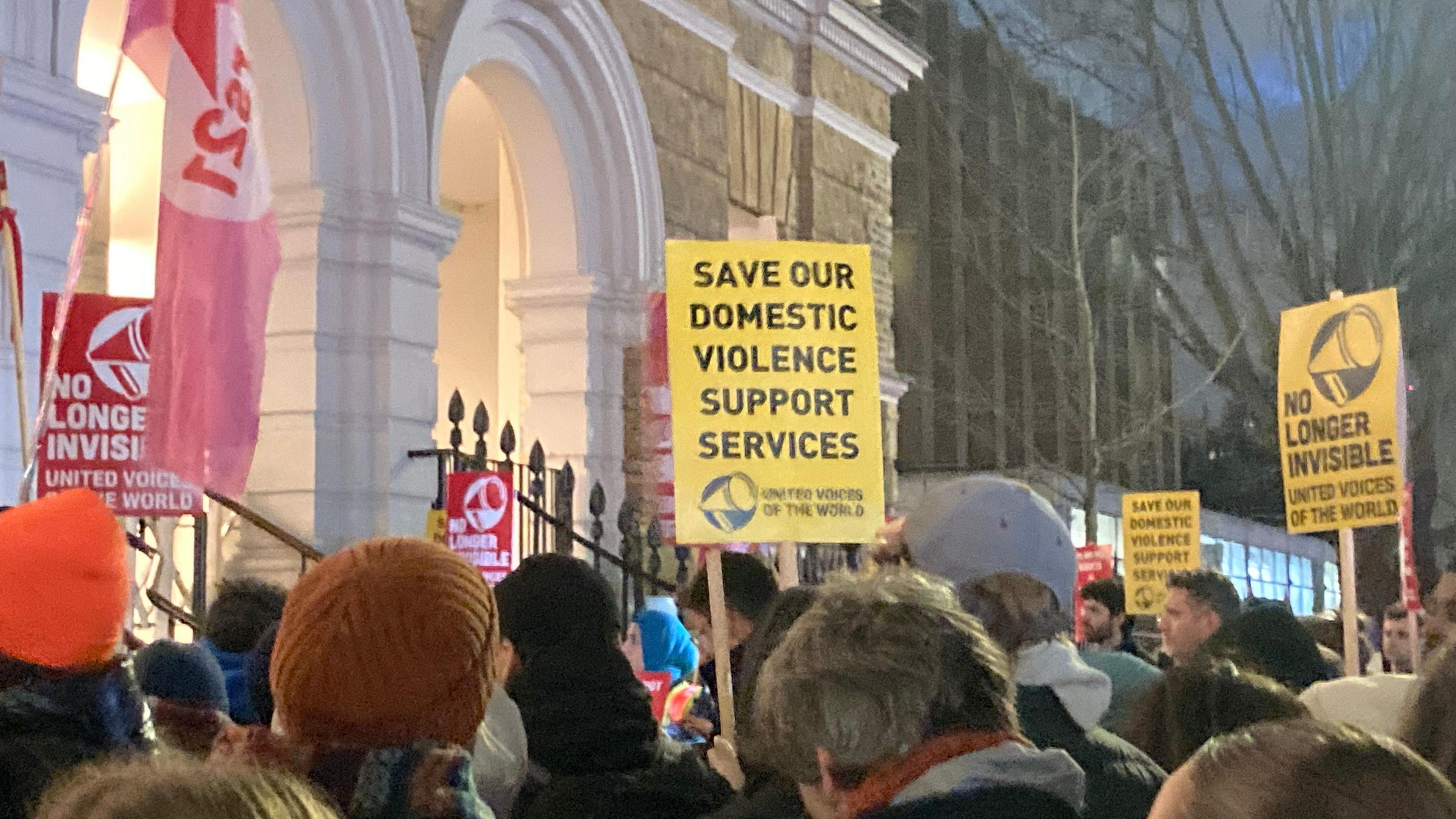 Protesters hold signs including 'save our domestic violence support services' and 'no longer invisible' outside a brick building with white arches