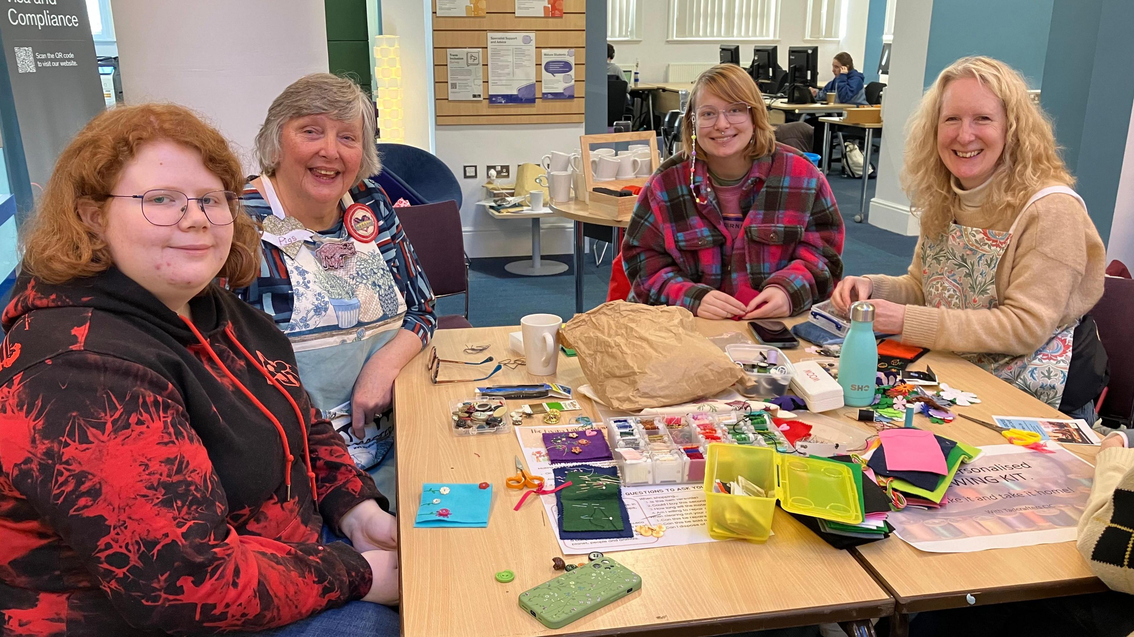 Two members of the Tadcrafters are sat at a table with two students, teaching them sewing skills. Sewing equipment including thread and pairs of scissors is spread across the table.