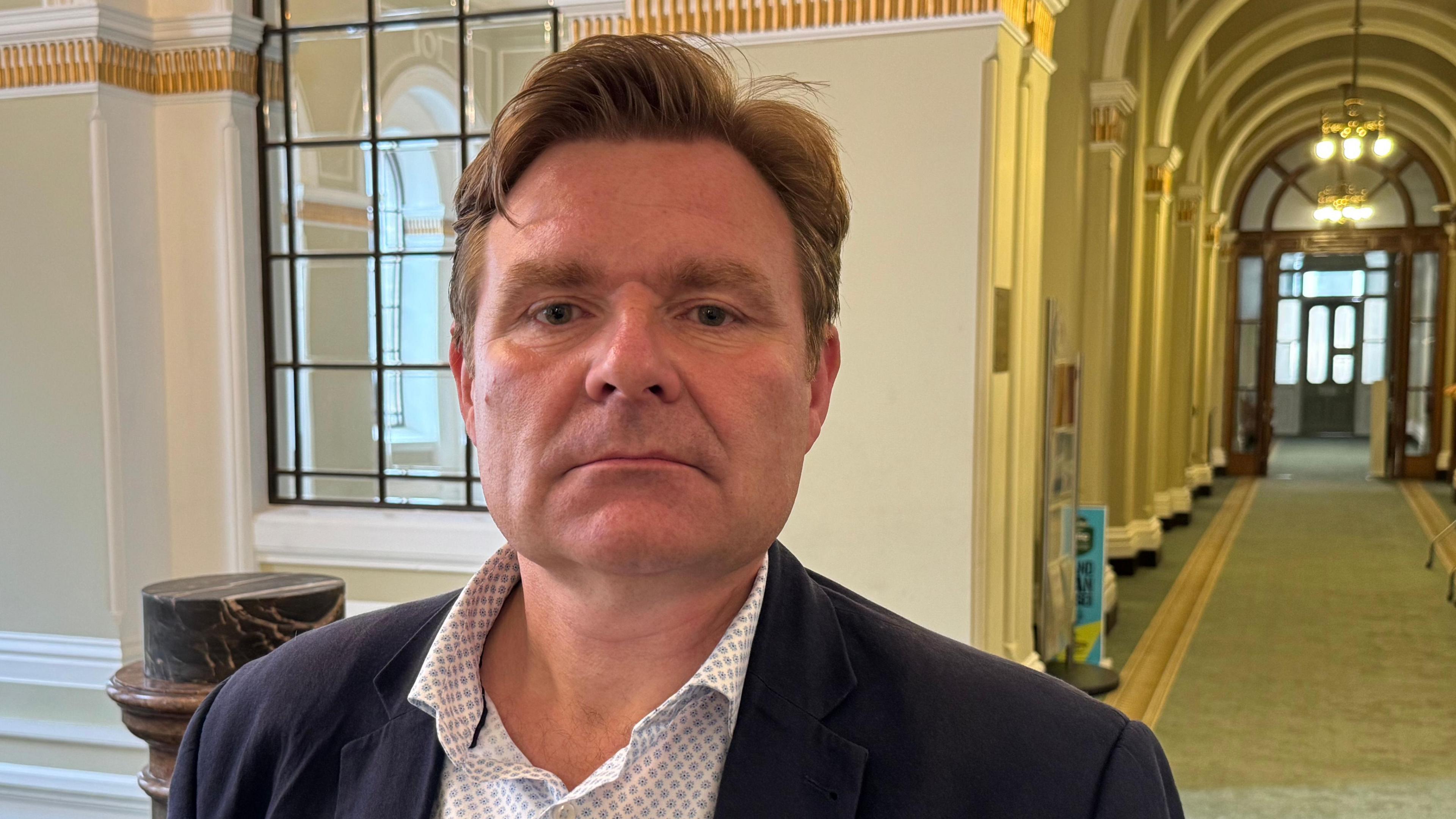 John Cotton standing in a council building wearing a shirt and jacket and looking serious.