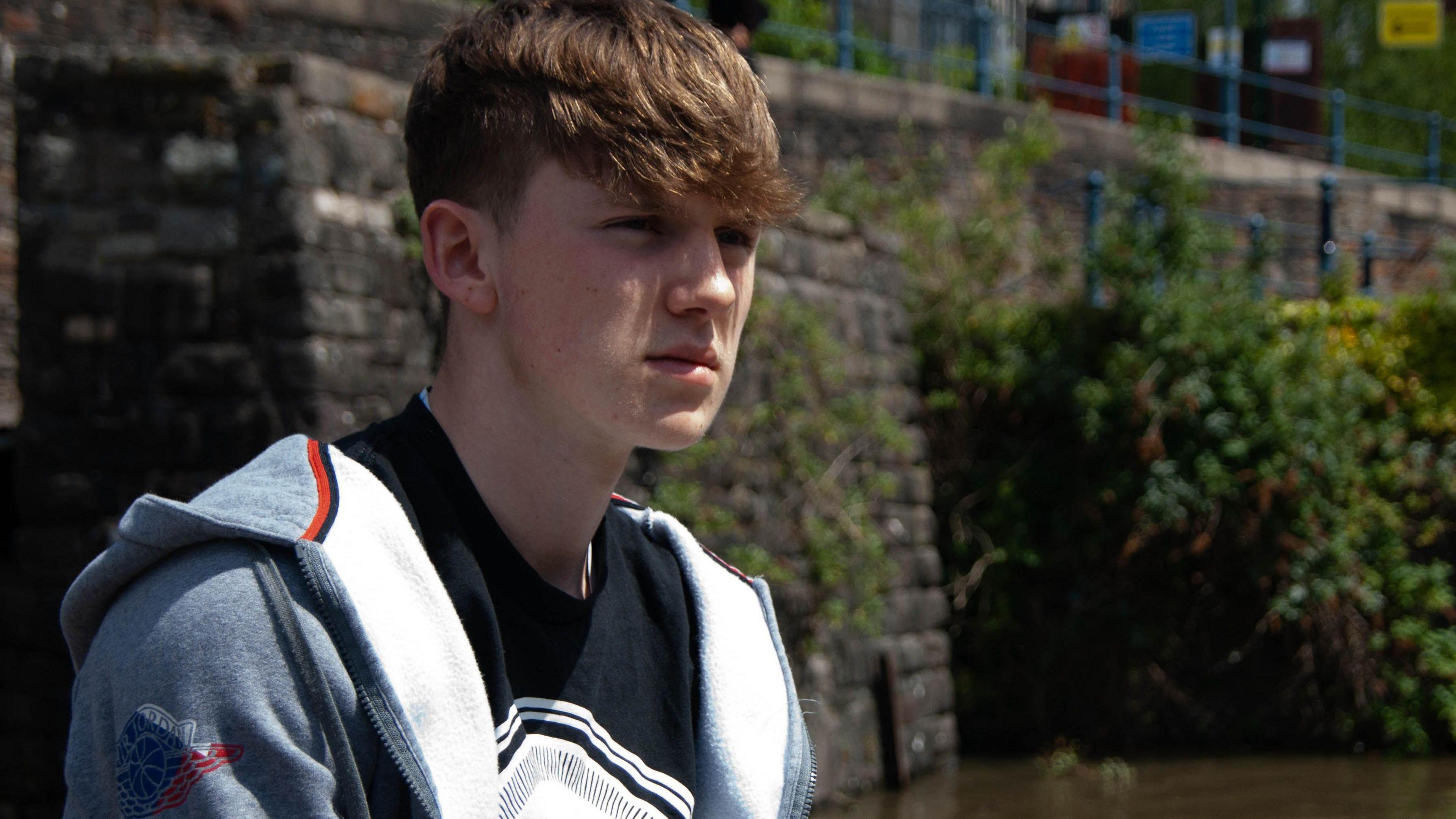 Calum Leighton pictured by the harbourside in Bristol. He is wearing a black t-shirt with a white image printed on it and a grey hooded top which is unzipped.