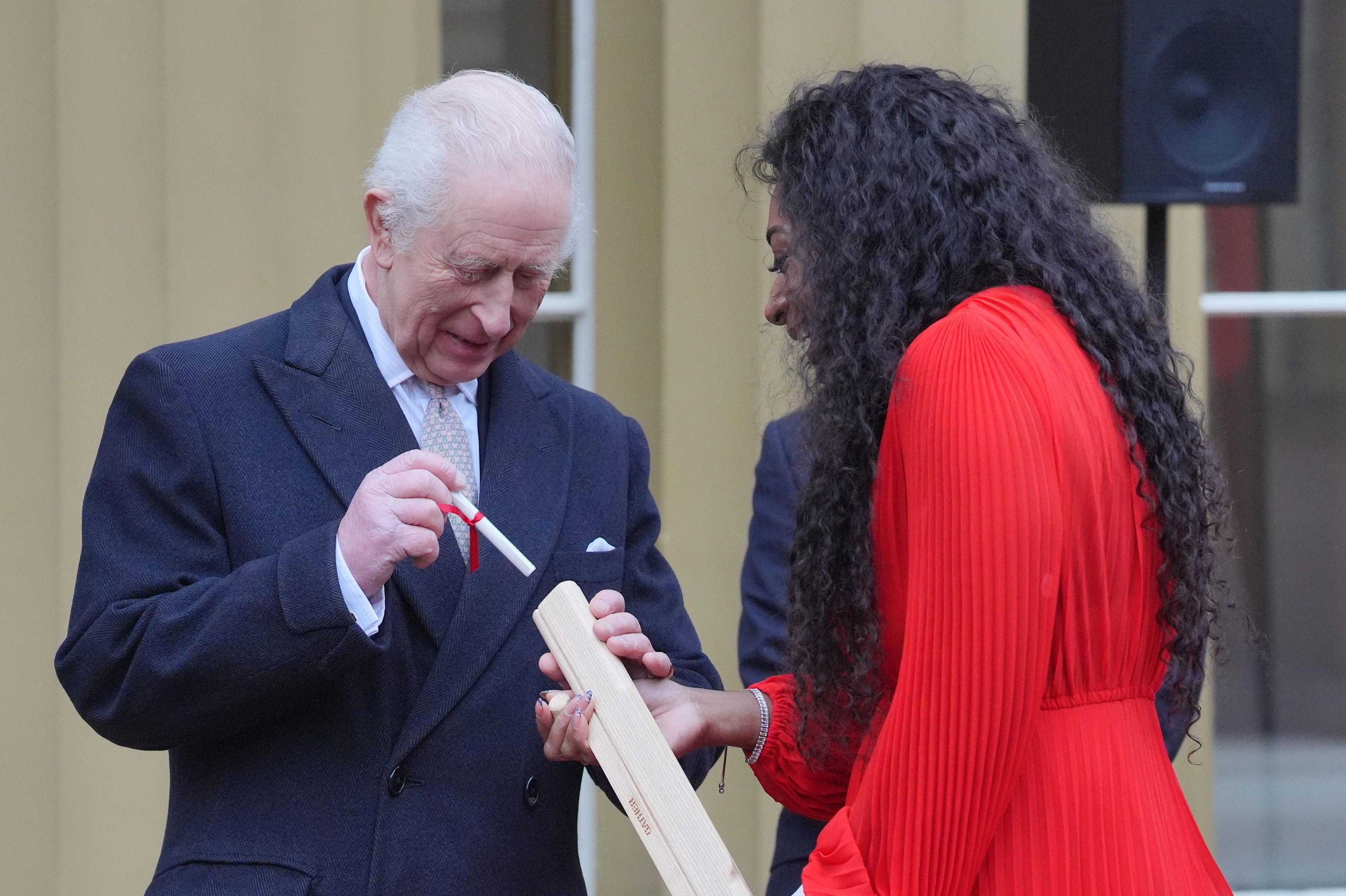 King Charles III in a suit places a rolled up piece of paper tied with a red ribbon into a golden baton held by parasport athlete Kadeena Cox