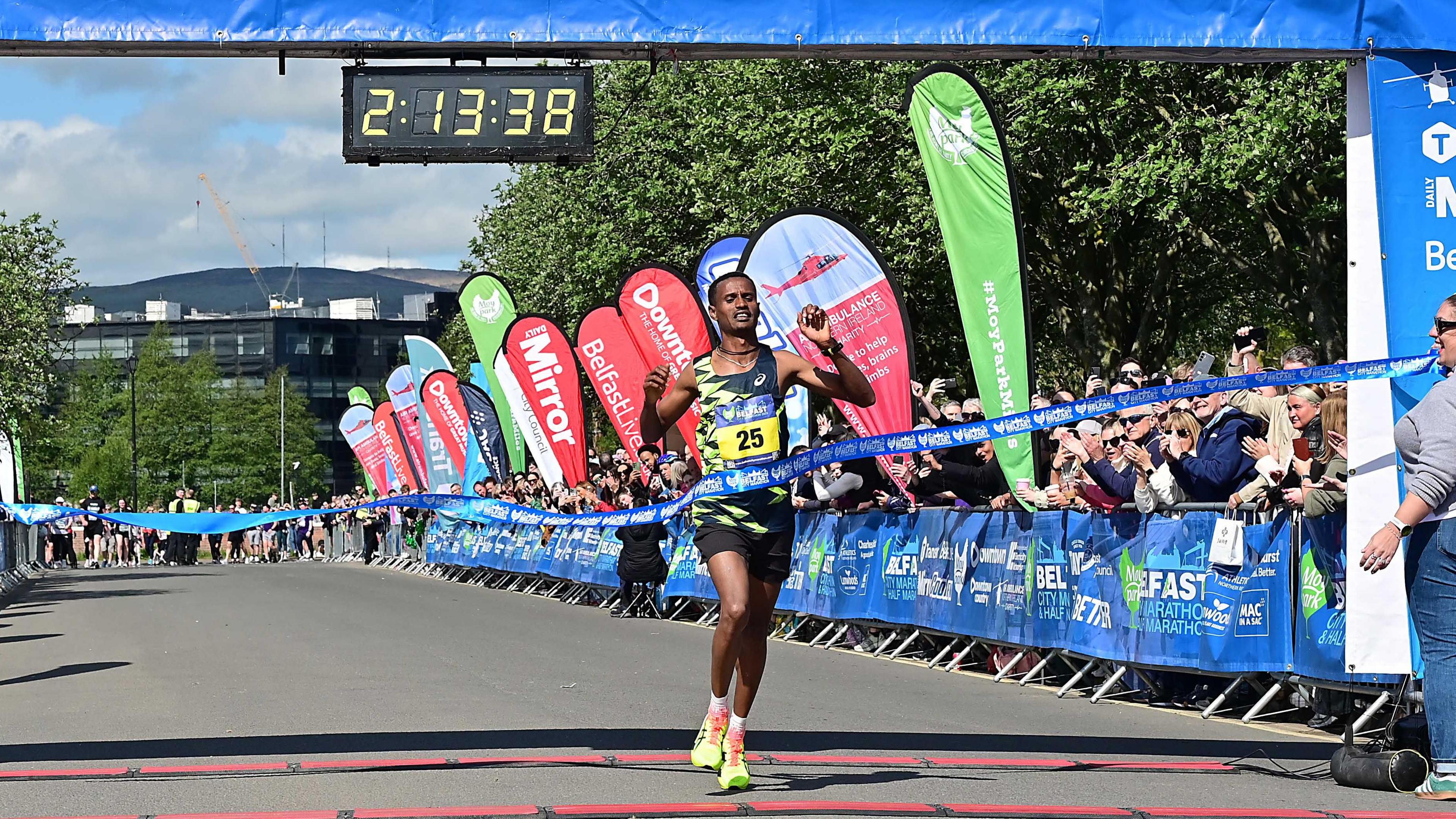 A man with short black hair crossing the finish line. He is wearing a black and yellow running vest, black shorts and yellow trainers. He is crossing through a blue ribbon and you can see the crowd cheering him on from behind a blue branded baricade.