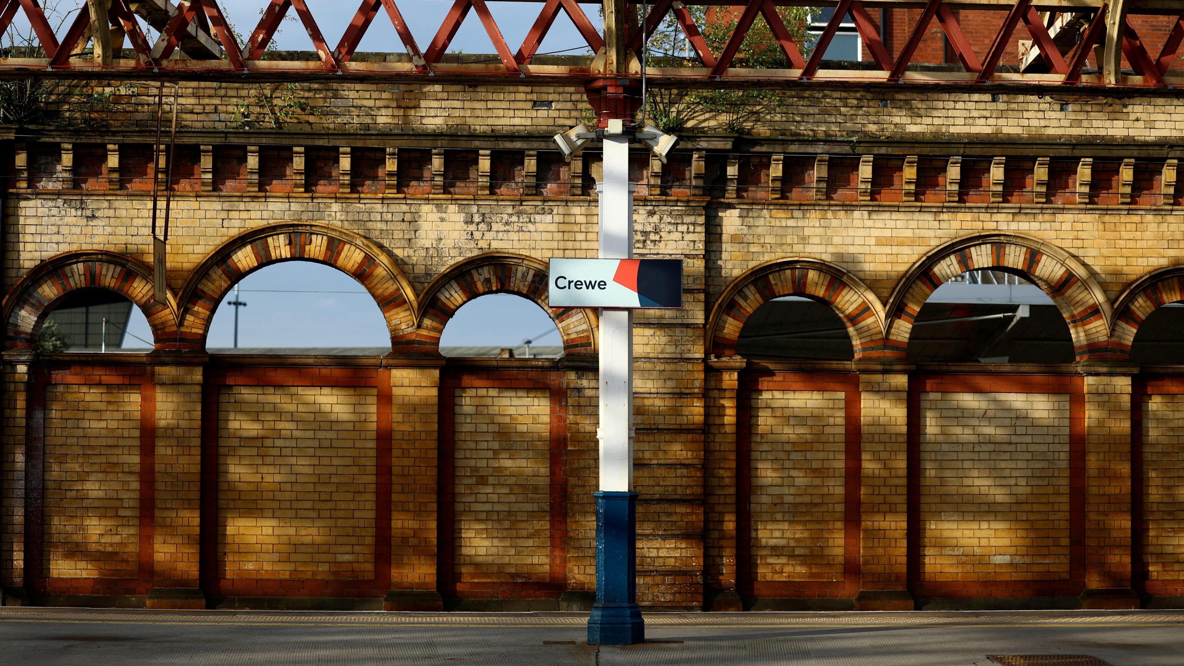 A railway platform with a brick wall of brown and orange bricks at the side of the platform, including a series of arches which are open to show the outside world of a blue sky. A metal post stands in the centre of the platform, painted white with a blue stump at the bottom. On the post is a sign with "Crewe" written on it.