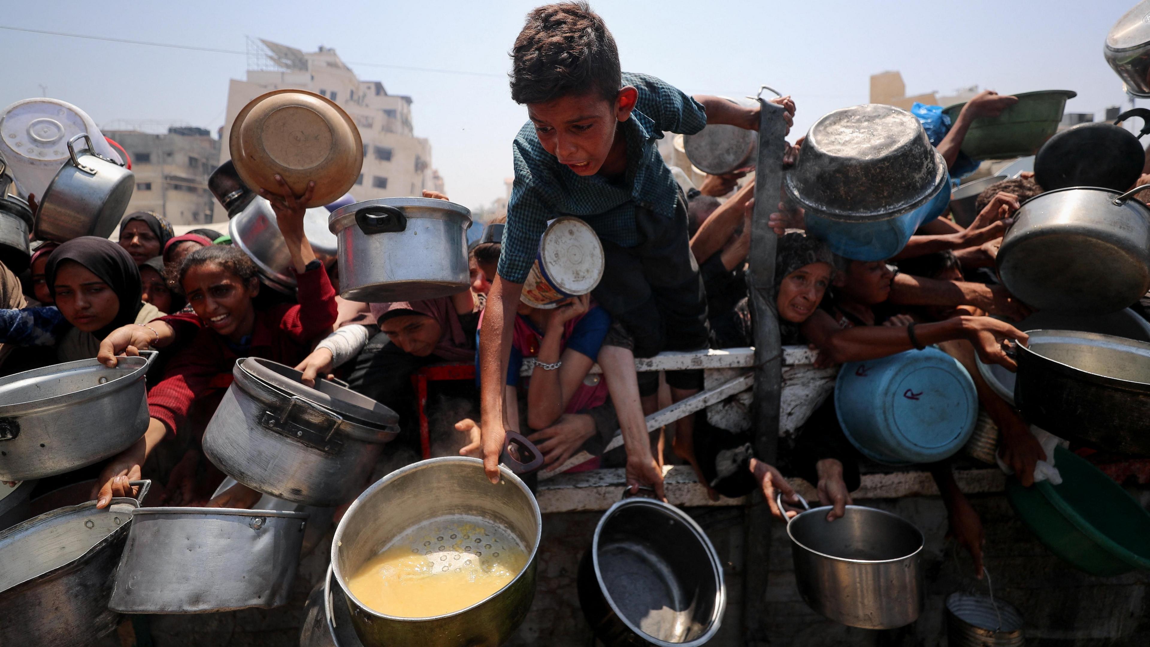 A frenzy of people hold saucepans and pots in front of the camera, in Gaza City. A little boy in a blue shirt leans far over the barrier holding people back, and appears to be shouting something.