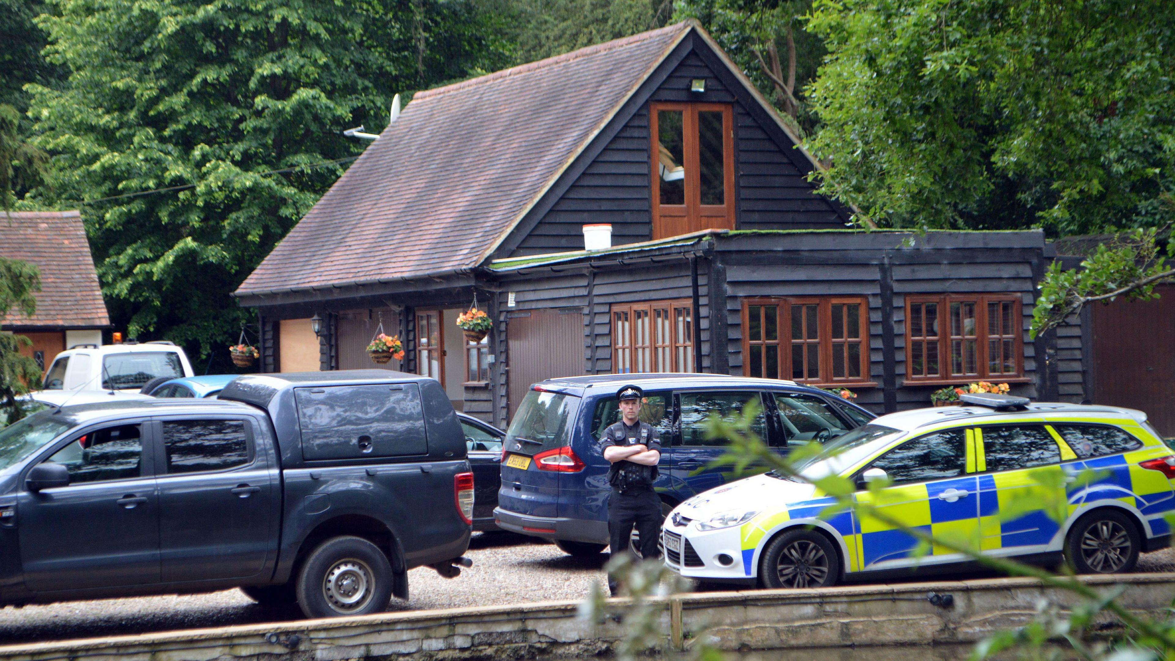 A uniformed police officer stands with his arms crossed beside a marked car, and surrounded by various other cars on the driveway area of John Palmer's home in South Weald. The picture has been taken a fair distance from the property.