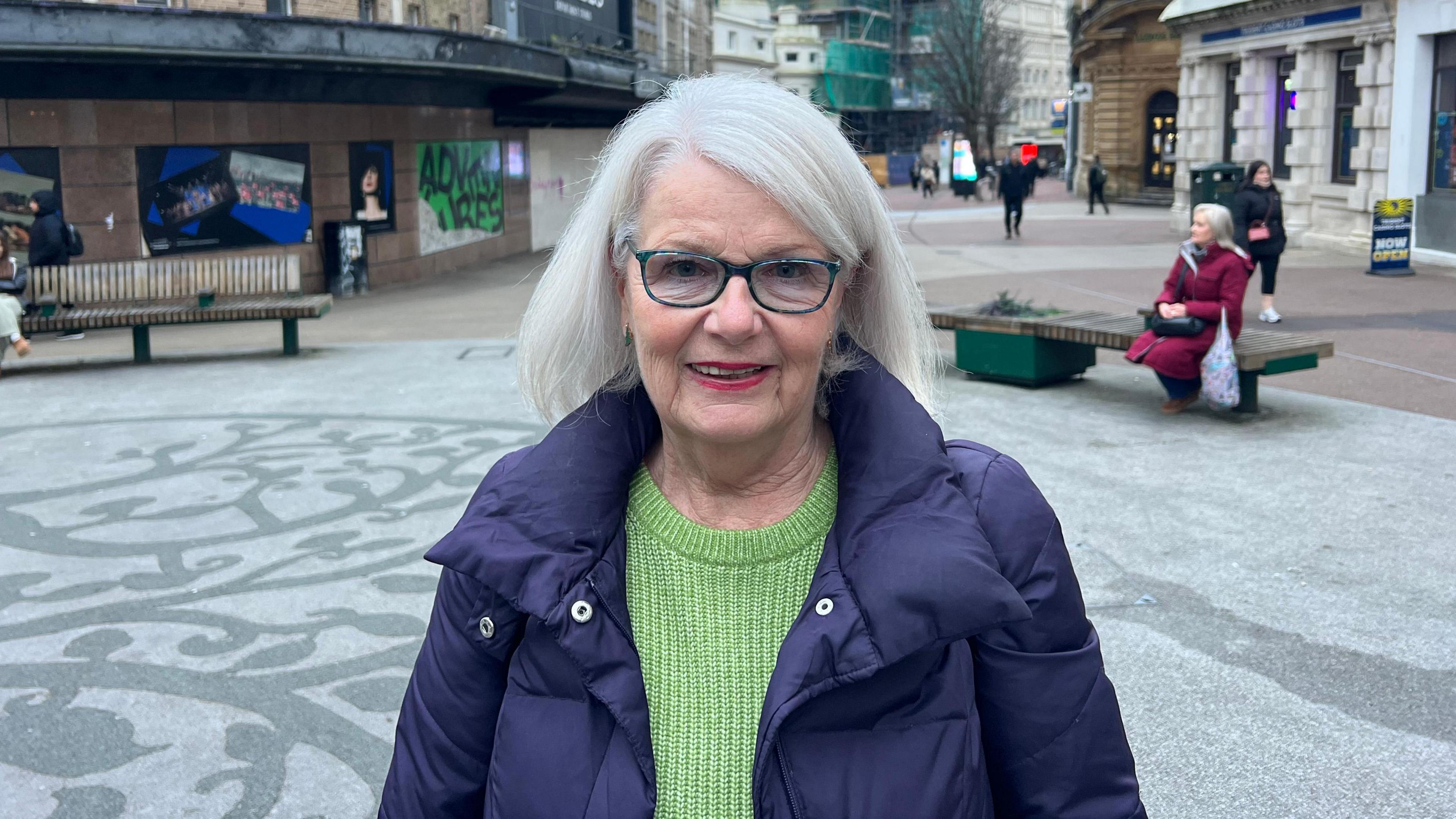 A woman stood in front of an empty shop, wearing glasses, a purple coat and green jumper.