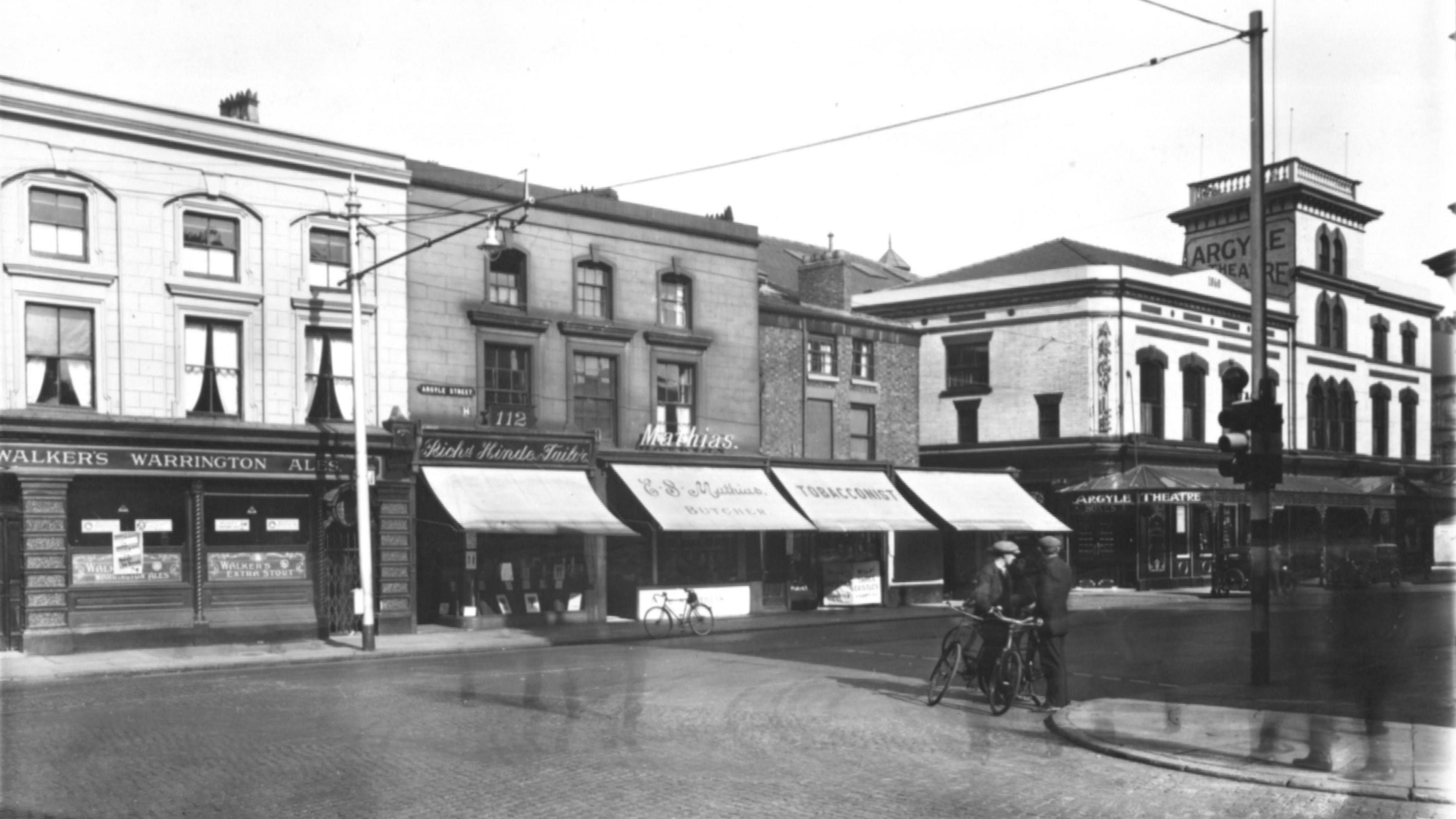 A black and white photo taken on Argyle Street showing the former theatre, a butchers, tobacconist, and an ale store. Two men wearing suits and flat caps can be seen talking on the corner, perched on their bicycles.
