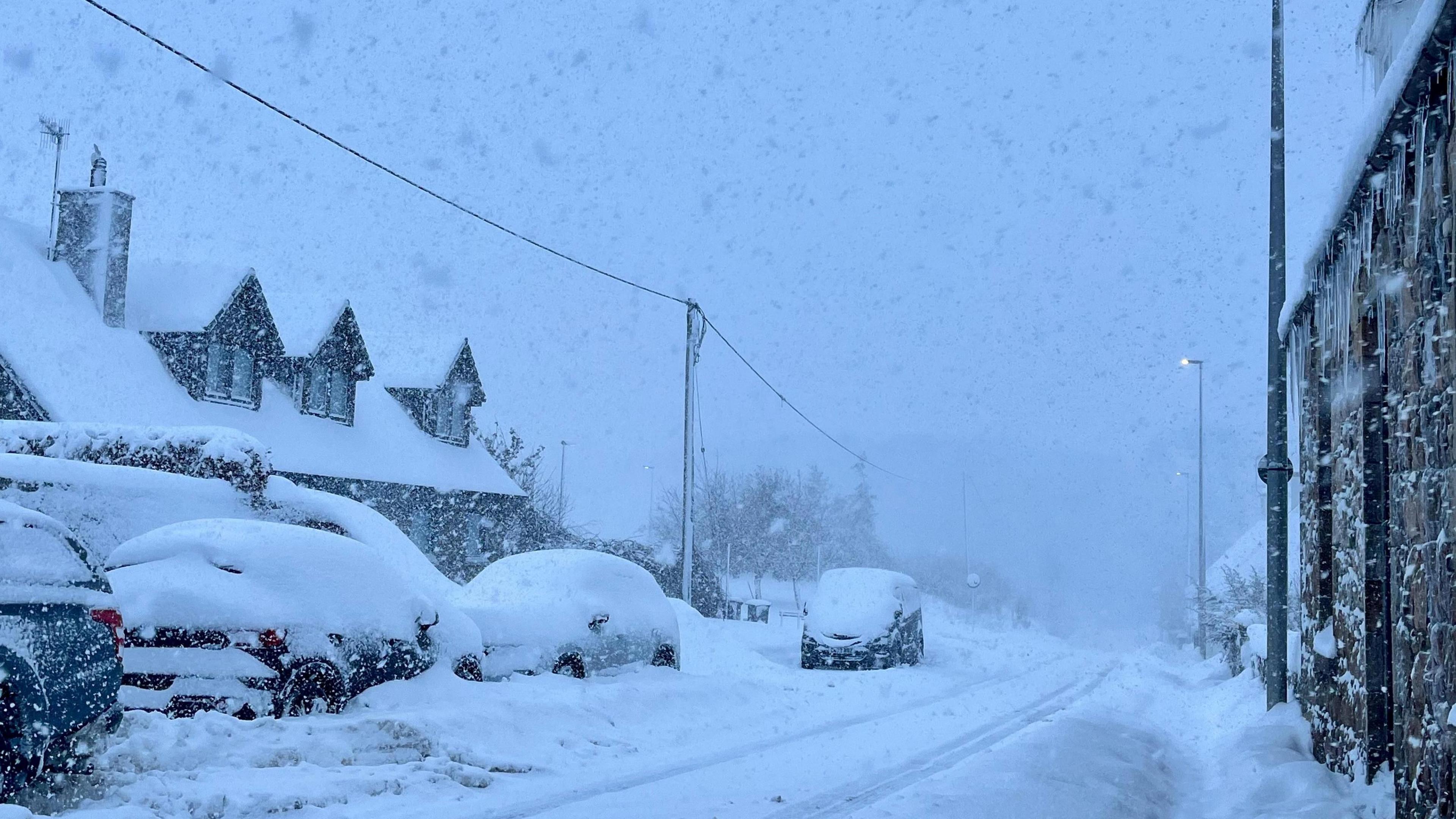 Street scene, with road, cars and houses covered in thick snow. Heavy snow is falling