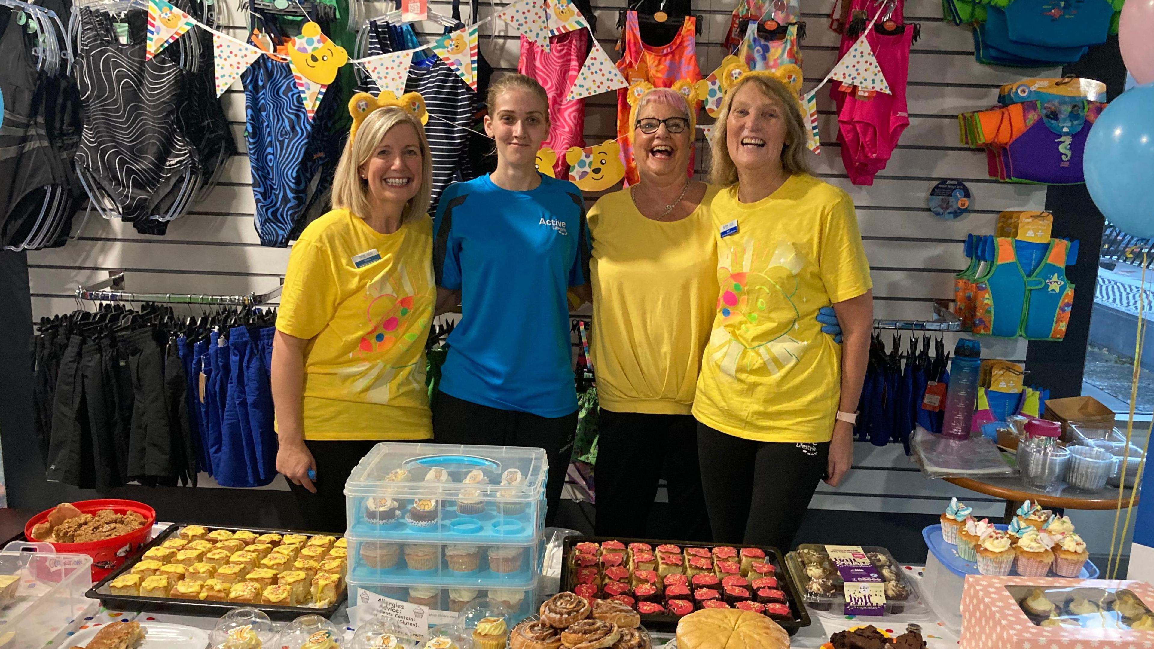 Four women stood behind a table full of cakes and baked goods. Three of them are wearing yellow T-shirts and yellow Pudsey ears, while one wears a blue T-shirt and no ears. They are all looking at the camera and smiling.