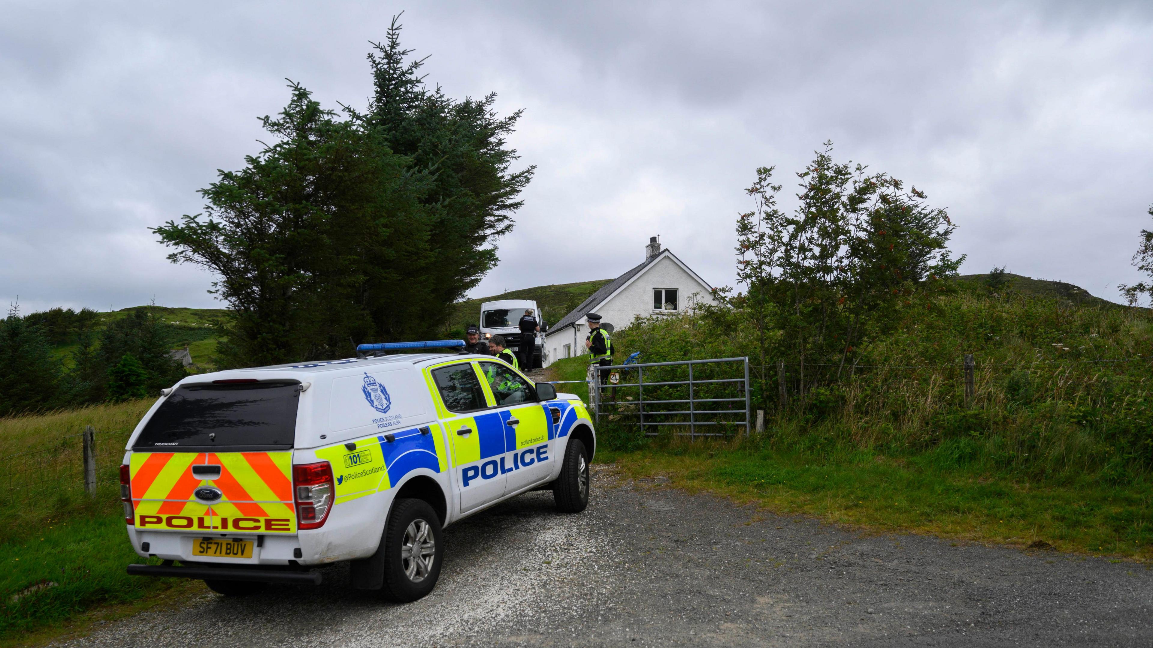 A Police car outside a house on Skye. The police car is long and white with orange and yellow striped on the back and the word "police" in large orange letters above the number plate. On the side is the blue Police Scotland logo above yellow and blue squares which cover the lower part of the car. The word "police" is on the side of the doors in blue. The house in the background is surrounded by greenery and is white with a dark roof. At least three police officers can be seen behind the vehicles and a white van is also parked outside the house.