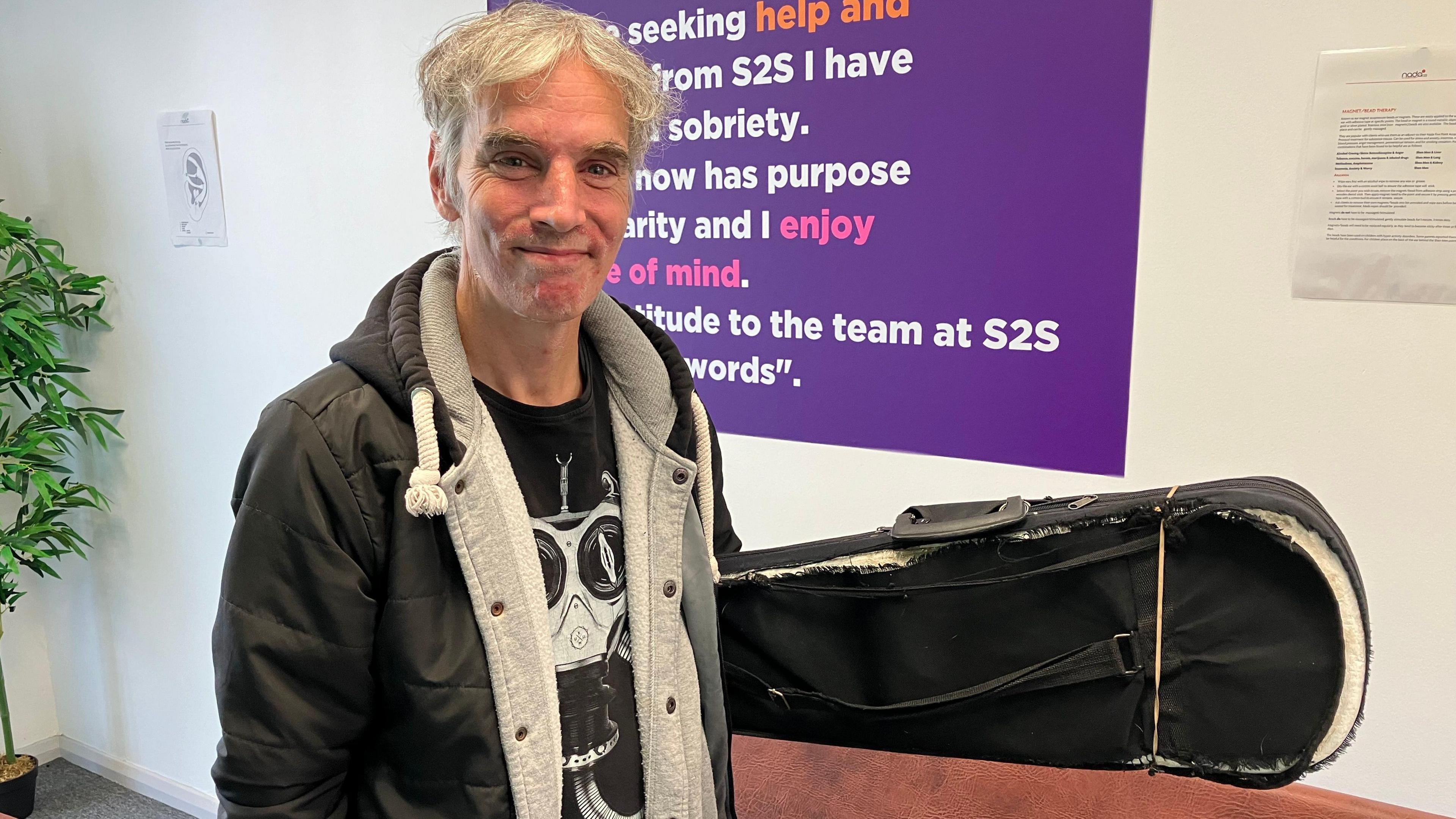 A man wearing a black hoodie and black t-shirt holding a violin case which is extremely frayed and held together with an elastic band.