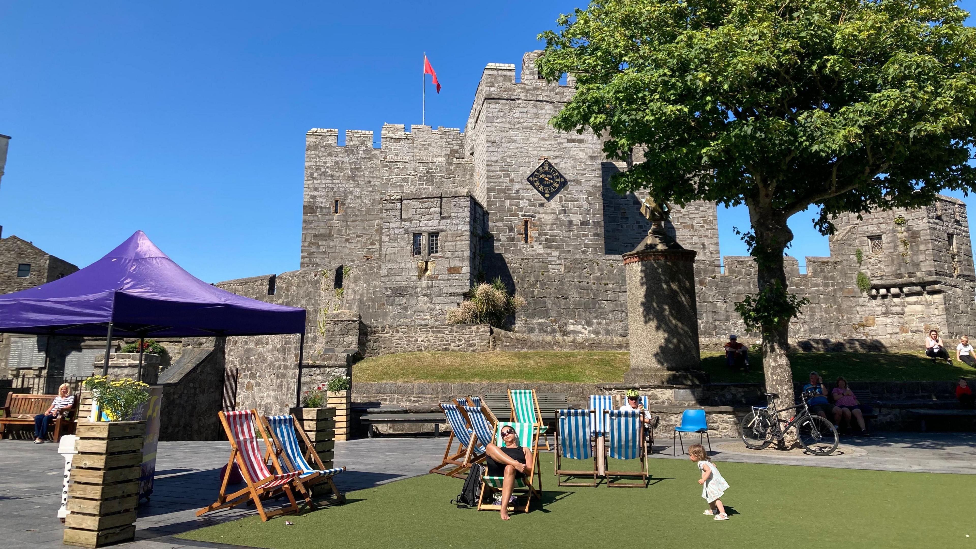Deck chairs in the square there is astro on the ground and a purple tent with a piano, with a medieval castle in the backgrround.