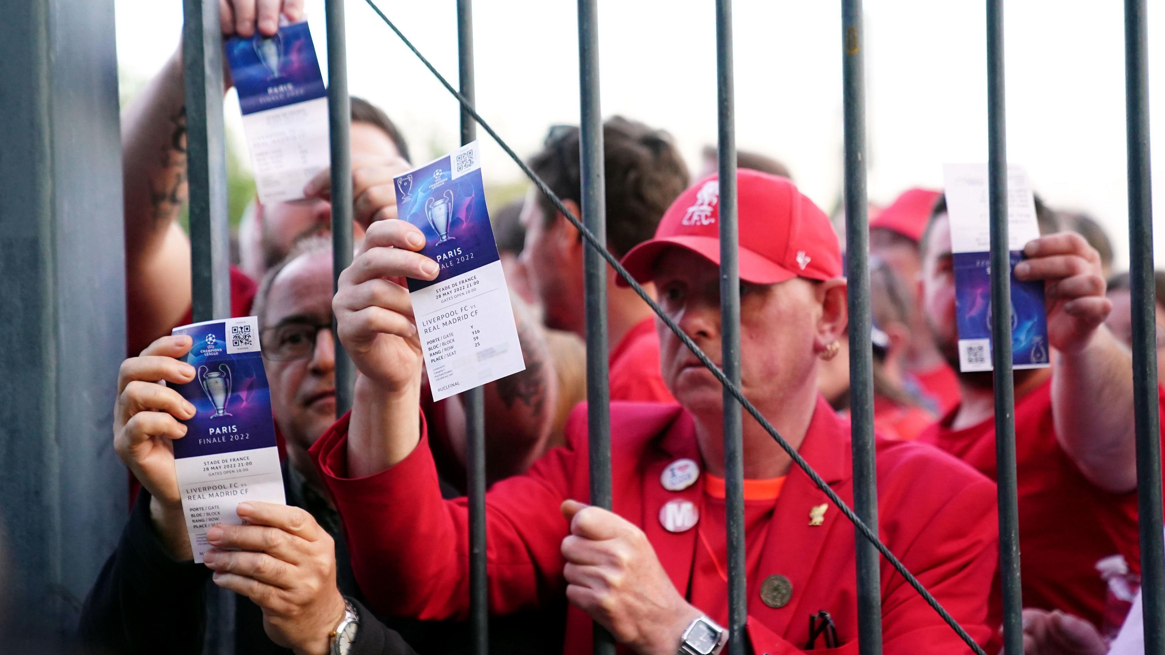 Liverpool fans clad in red shirts hold up their tickets to the Champions League 2022 final match in Paris from behind an iron gate