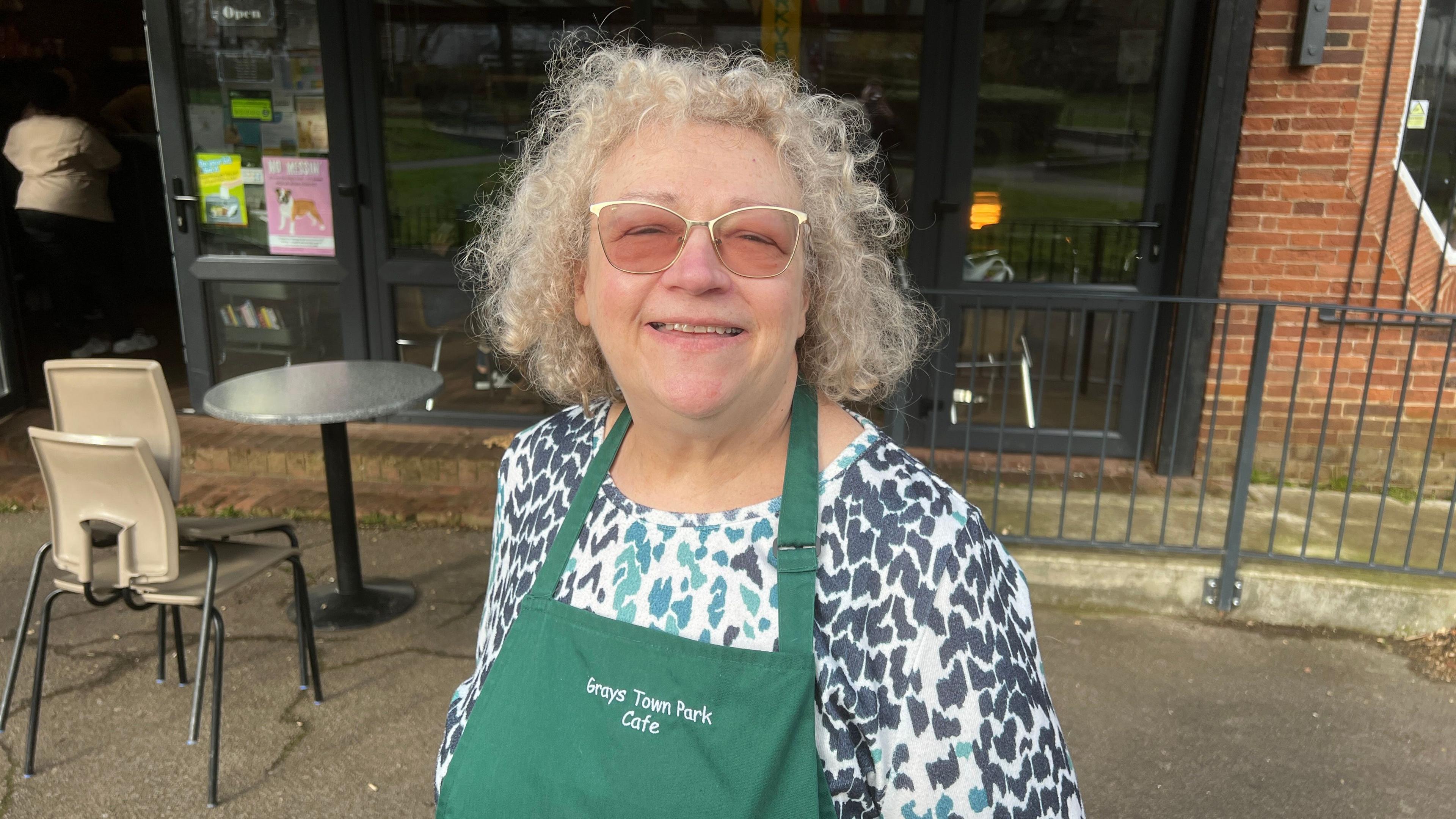 Gillian Rainbow wearing a navy blue leopard print top and wearing a green apron. She has curly blonde hair and is wearing sunglasses. She is standing outside in front of a cafe.