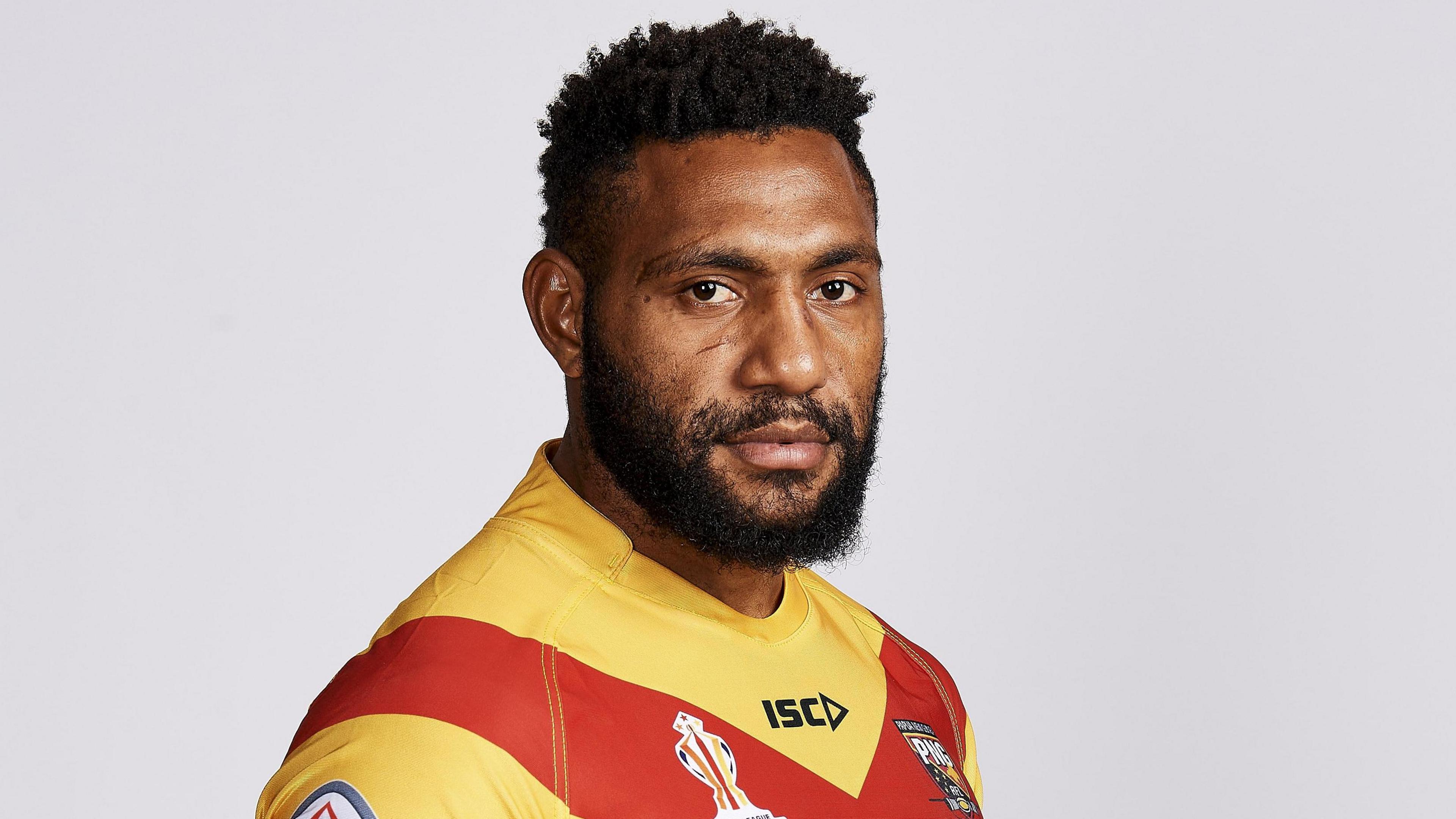 Rugby league player Keven Appo, wearing a yellow and red rugby shirt. Pictured side on against a grey backdrop, he is looking directly at the camera.