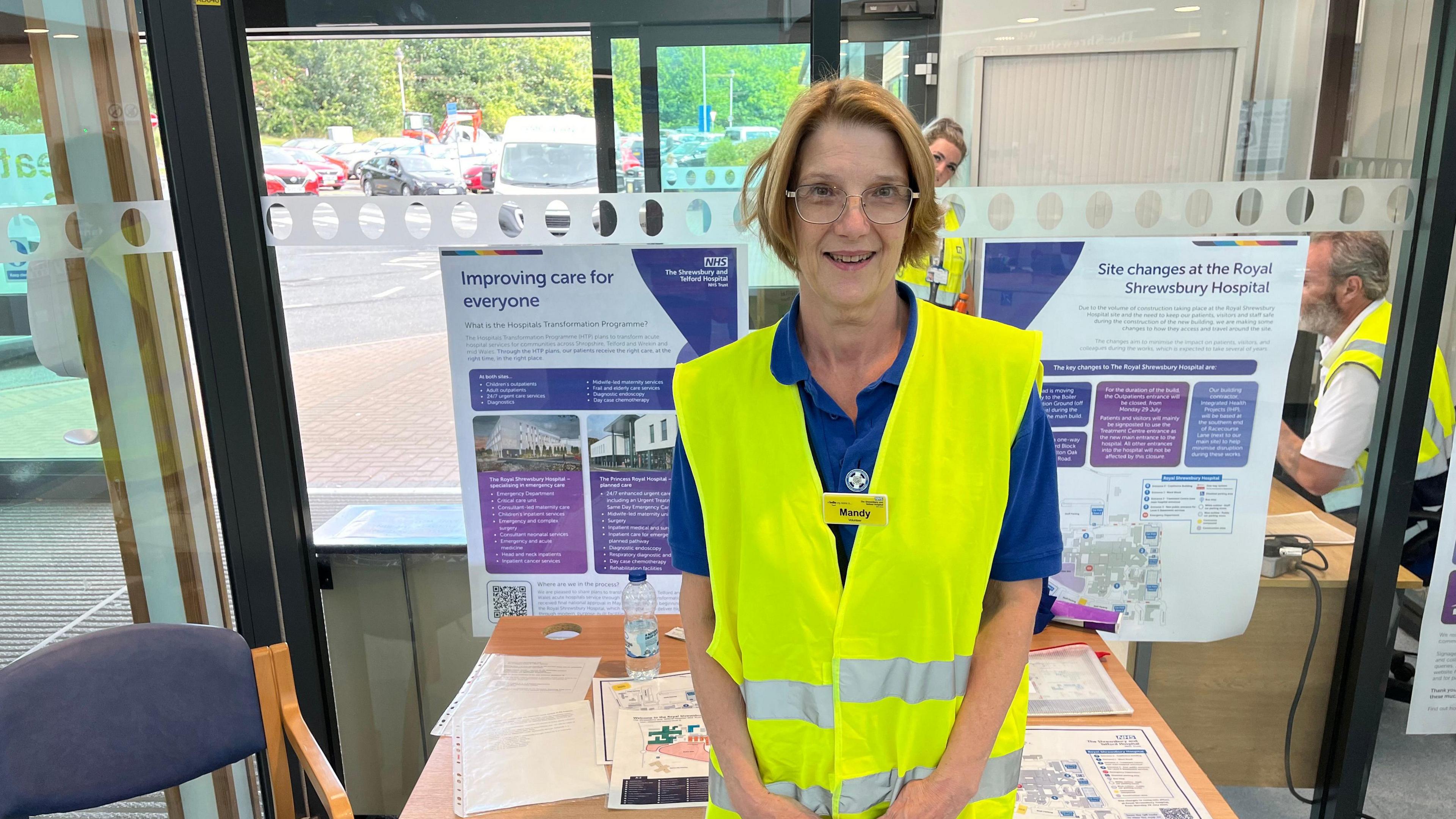 Mandy has short, strawberry blonde hair and is wearing glasses. She is standing in front of an information desk smiling. She is wearing a blue t-shirt and a yellow, hi-vis tabard.